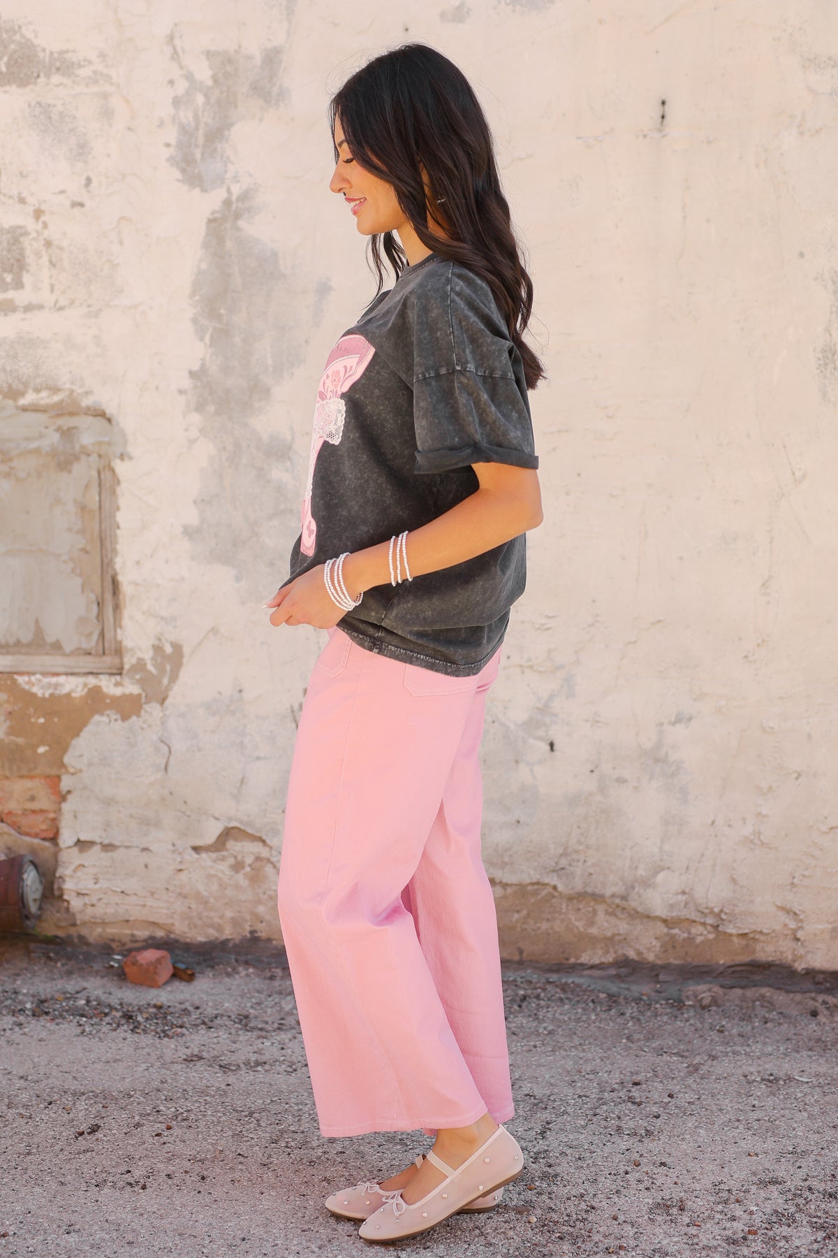 Woman wearing a dark gray t-shirt with a graphic design and light pink pants against a textured wall.