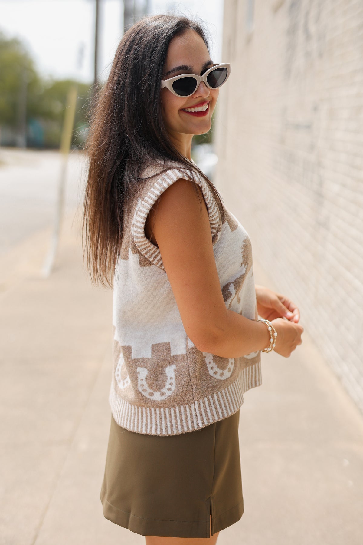 Brunette model, outside, wearing a light mocha sweater vest, tan boots, and a chocolate colored skort.