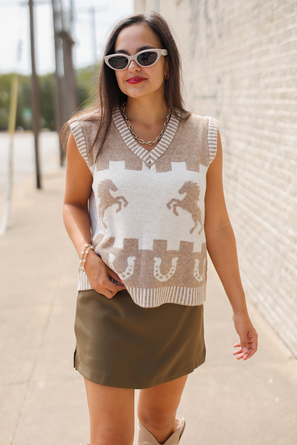Brunette model, outside, wearing a light mocha sweater vest, tan boots, and a chocolate colored skort.