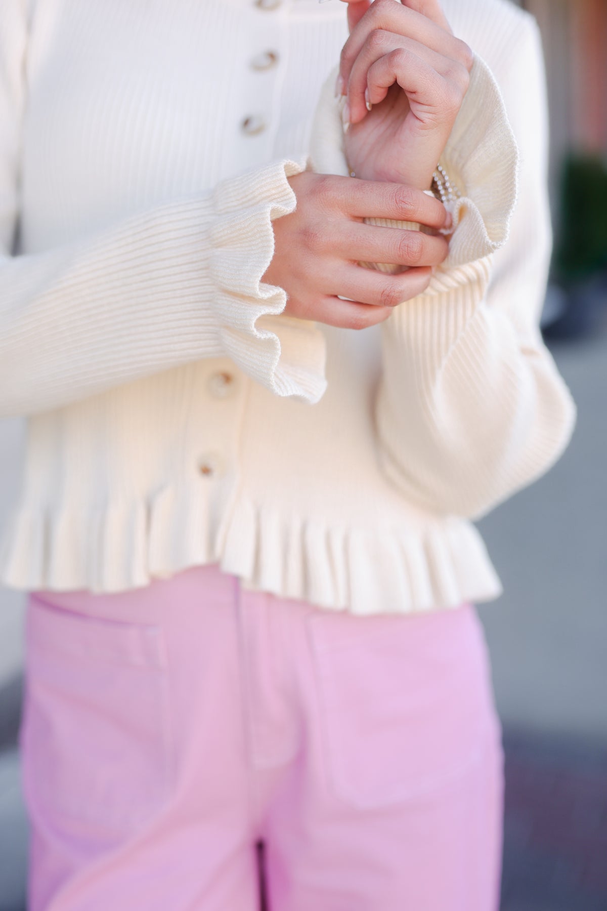 Close-up of a person wearing a white cardigan with ruffled sleeves and pink pants.