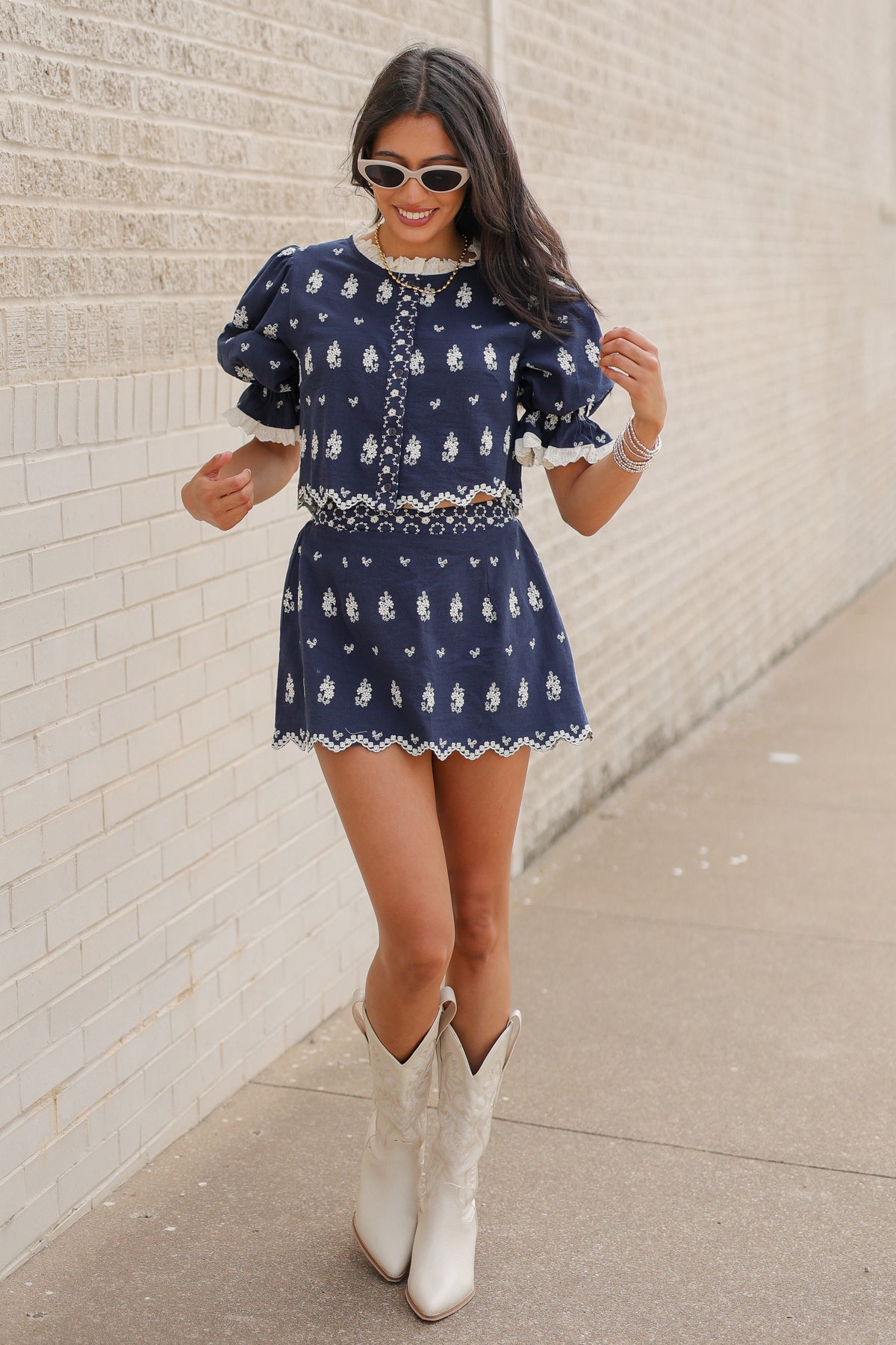 Brunette in a blue set standing outside wearing white cowboy boots and sunglasses. Southern House Clothing