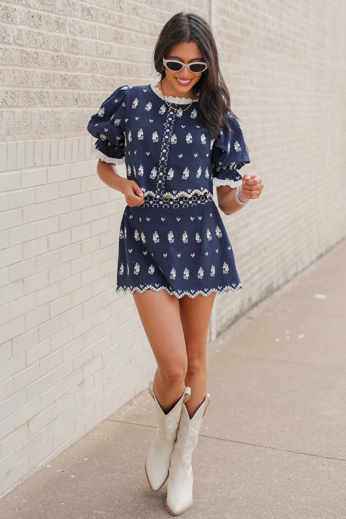 Brunette in a blue set standing outside wearing white cowboy boots and sunglasses. Southern House Clothing