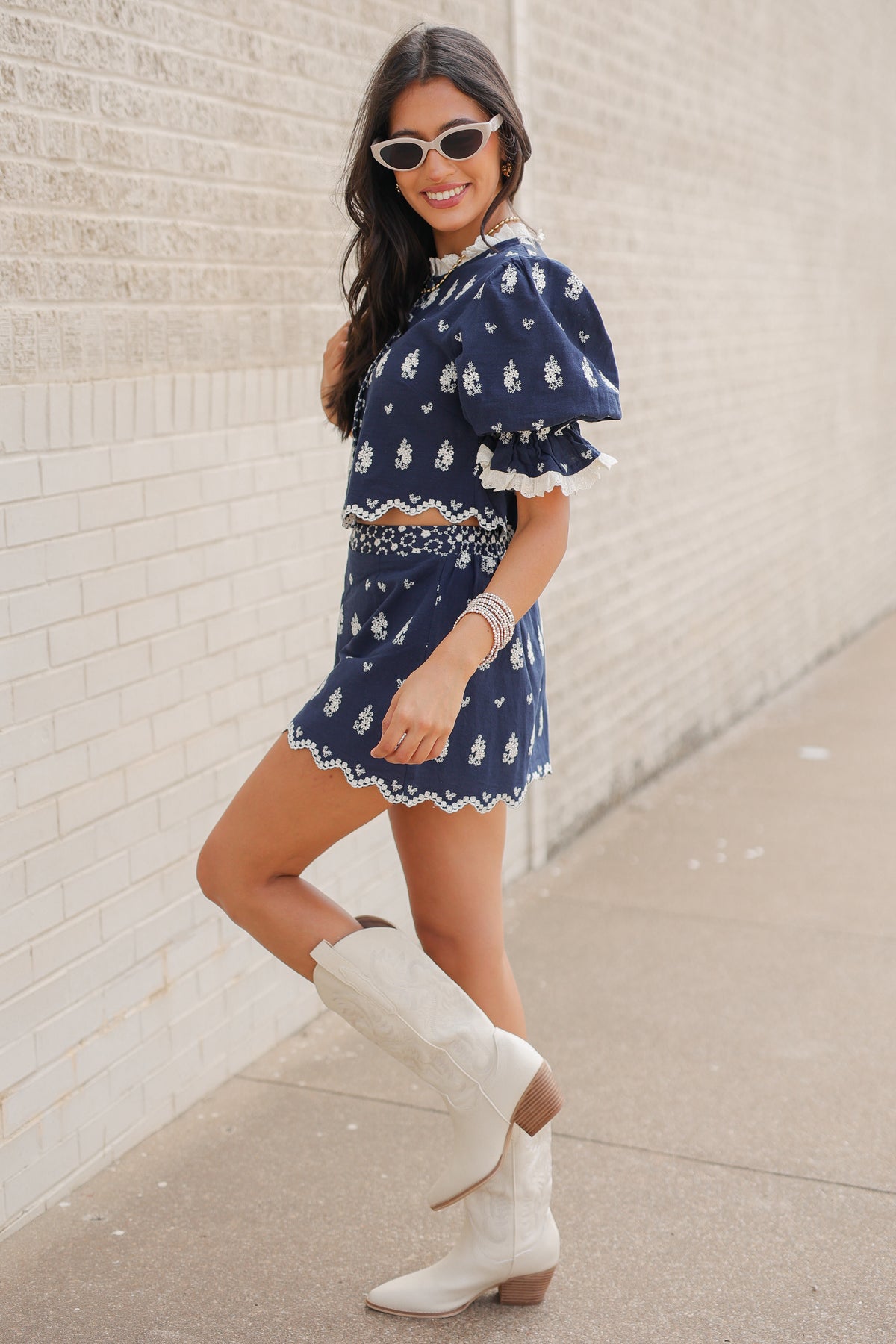 Brunette in a blue set standing outside wearing white cowboy boots and sunglasses. Southern House Clothing