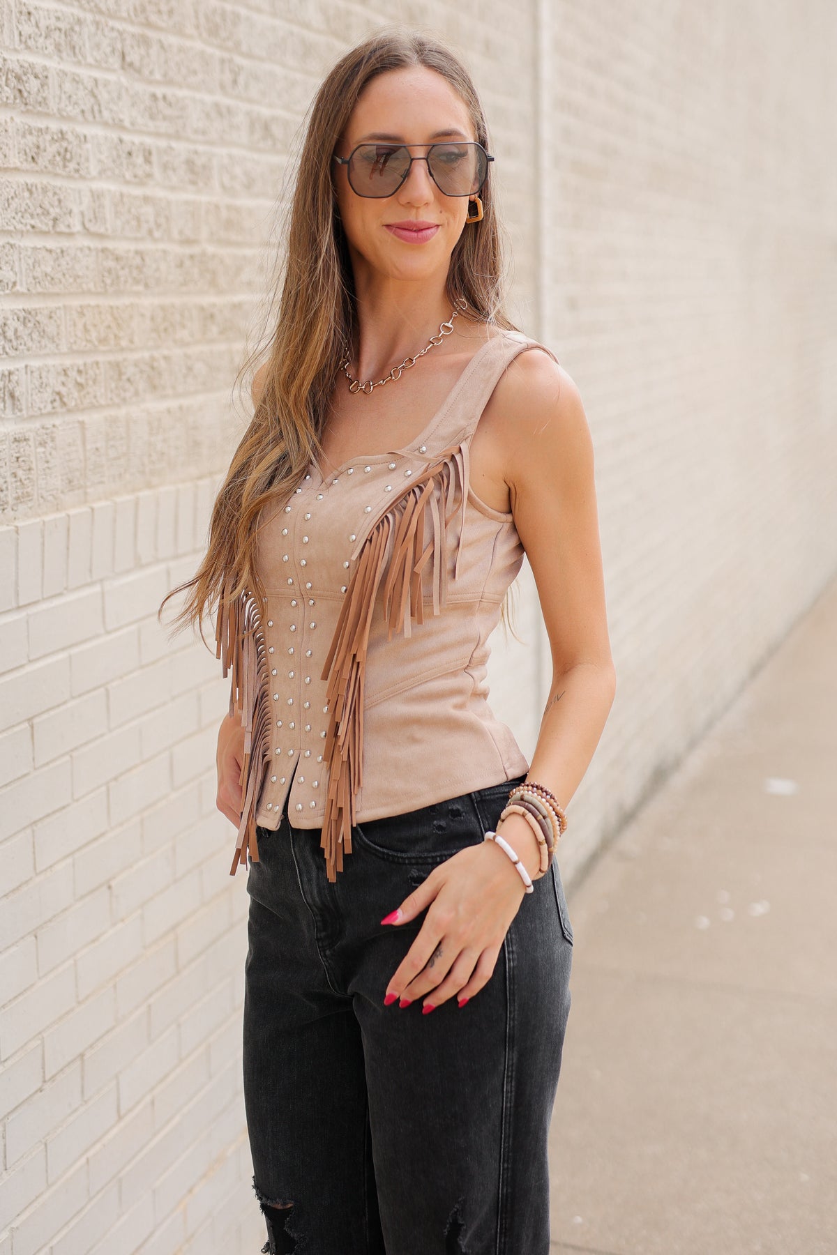 Blonde model wearing Laredo Luxe Studded Fringe Top in Camel featuring silver-tone studs, western fringe, sweetheart neckline, and wide straps, styled with black denim standing outside. Southern House Clothing