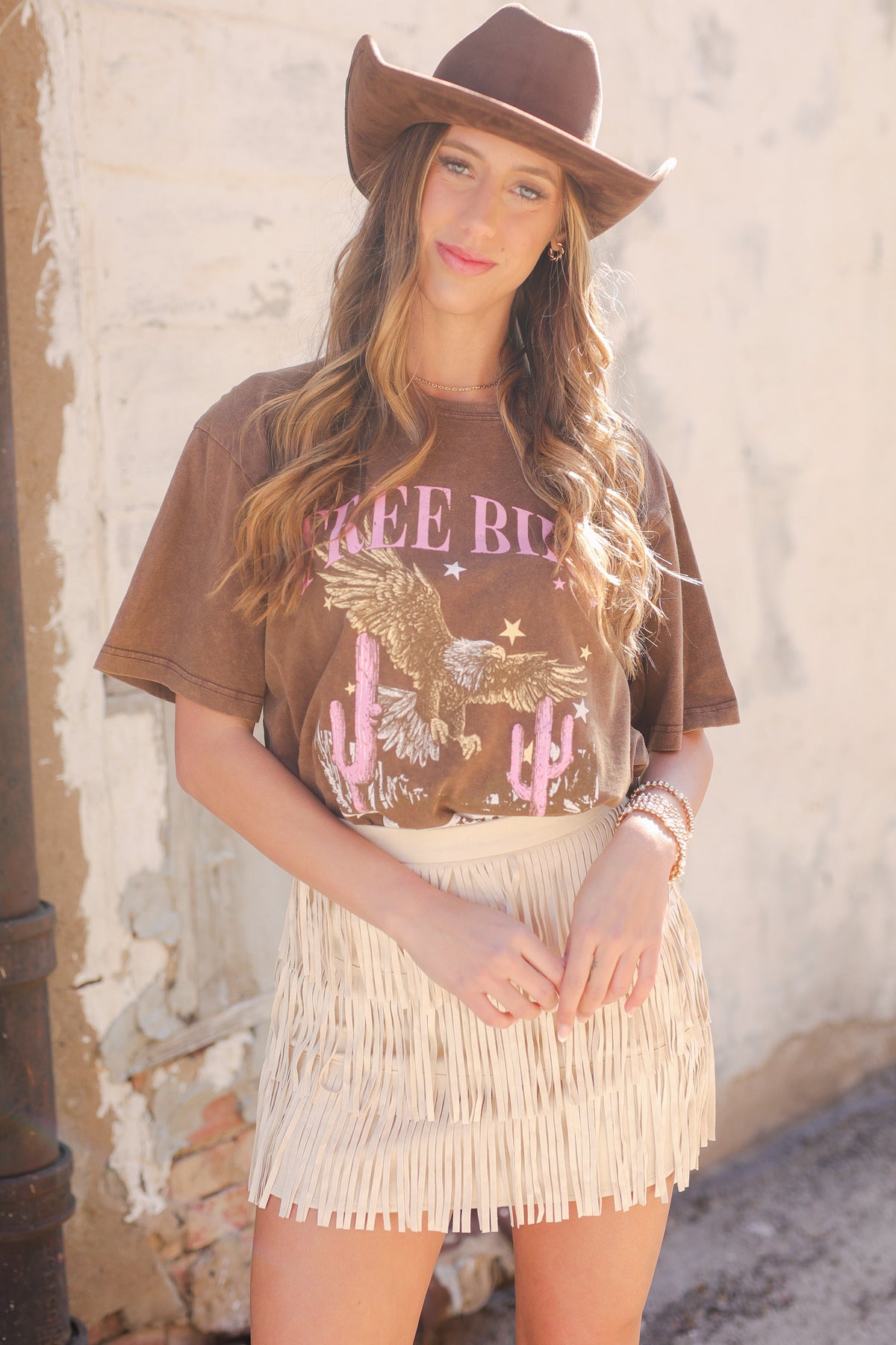 Woman wearing a brown graphic t-shirt and fringed skirt with a cowboy hat against a rustic wall.