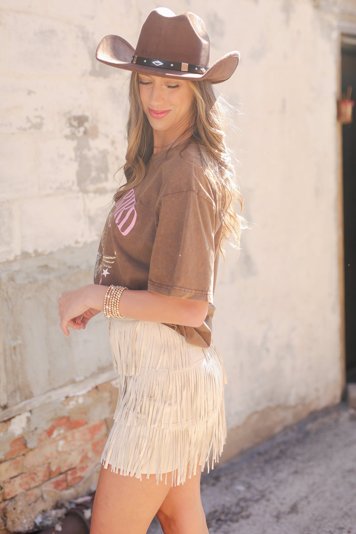 Woman wearing a brown cowboy hat and a fringe dress against a rustic wall.