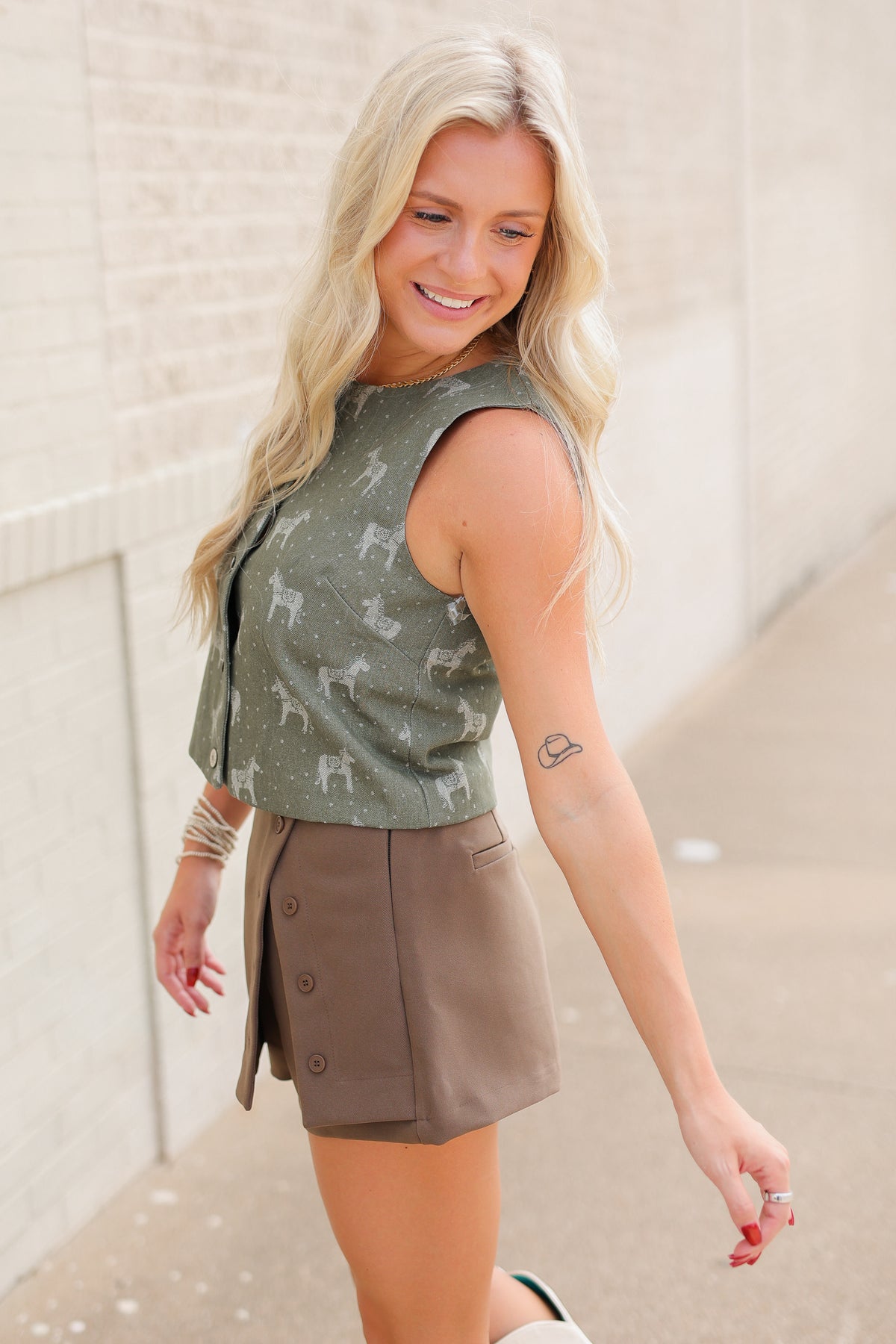 Blonde model wearing a green denim vest top with horse designs, a brown skort, and white cowboy boots - standing outside. Southern House Clothing