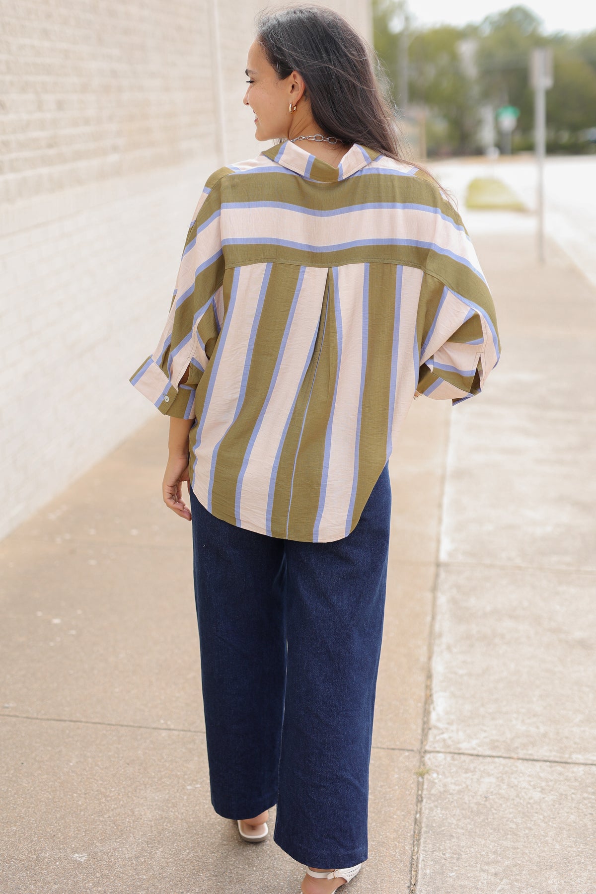 Back view of a model wearing Mabank Stripe Button-Up Top in olive green, ivory, and light blue with an oversized fit, collared neckline, and wide sleeves, styled with the Langley dark-wash jeans. Southern House Clothing