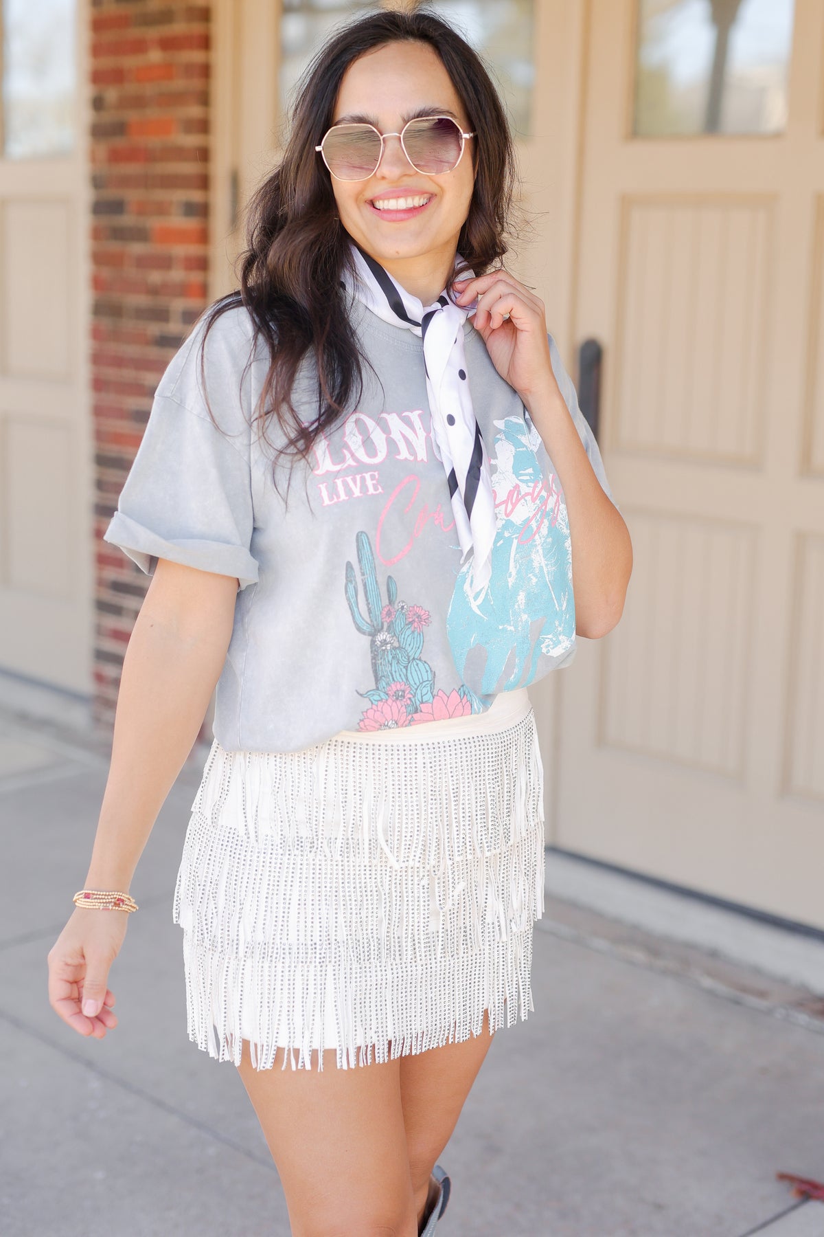 Woman wearing a gray t-shirt with colorful graphics and a white fringe skirt, standing outdoors.