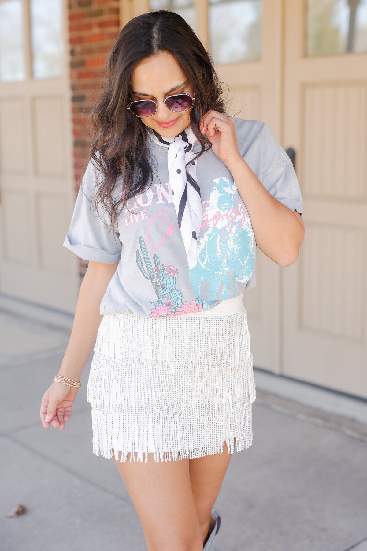Woman wearing a colorful blouse with a graphic design and a white fringe skirt, standing on a sidewalk.