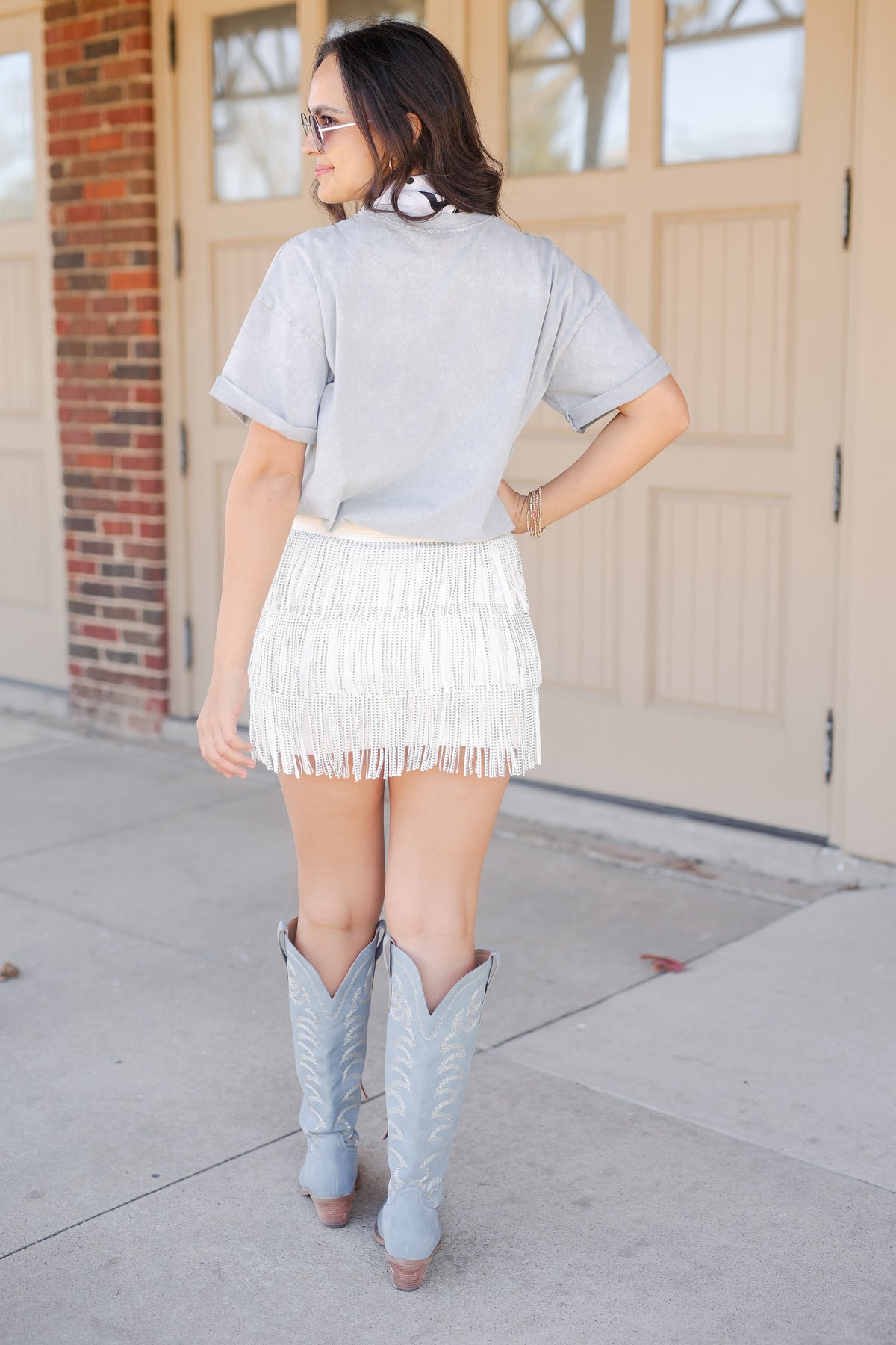 Woman wearing a light gray t-shirt, white fringe skirt, and gray cowboy boots standing on a sidewalk.