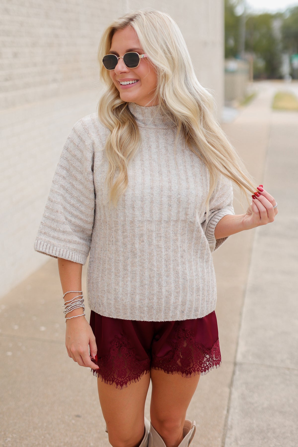Blonde model wearing an oatmeal color sweater top and burgundy laced trimmed shorts outside.