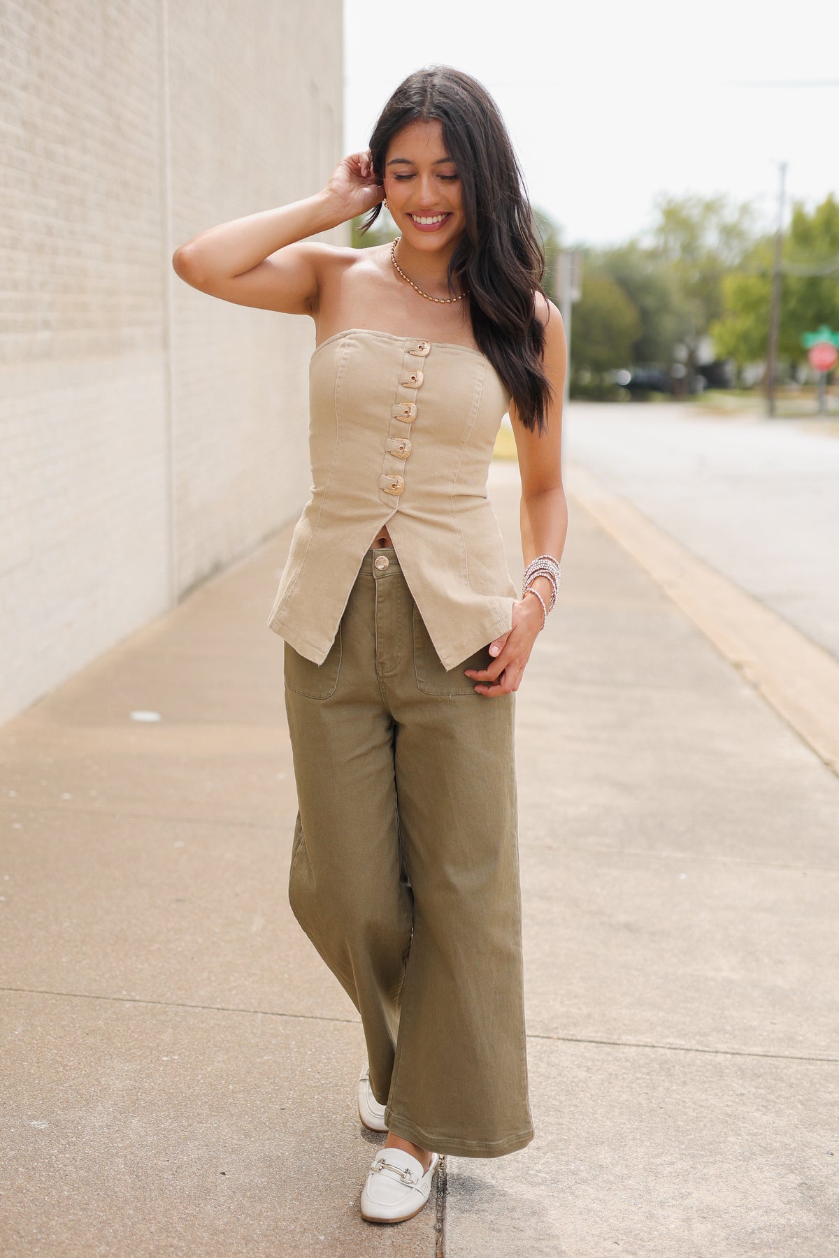 Brunette model wearing the Westlyn Buckle Strapless Top in Taupe featuring gold-tone buckle accents and a split-front hem, styled with olive wide-leg pants and white loafers standing outside. Southern House Clothing.