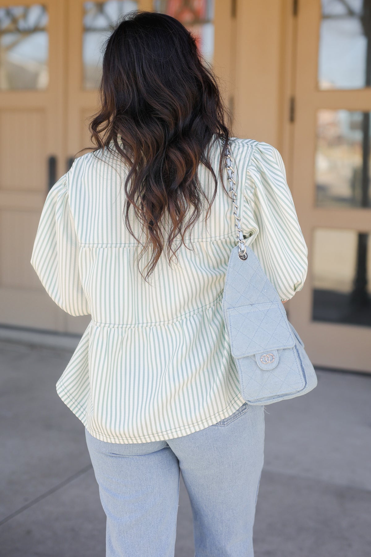 Woman wearing a light-colored striped blouse and light blue jeans, standing in front of a glass door.