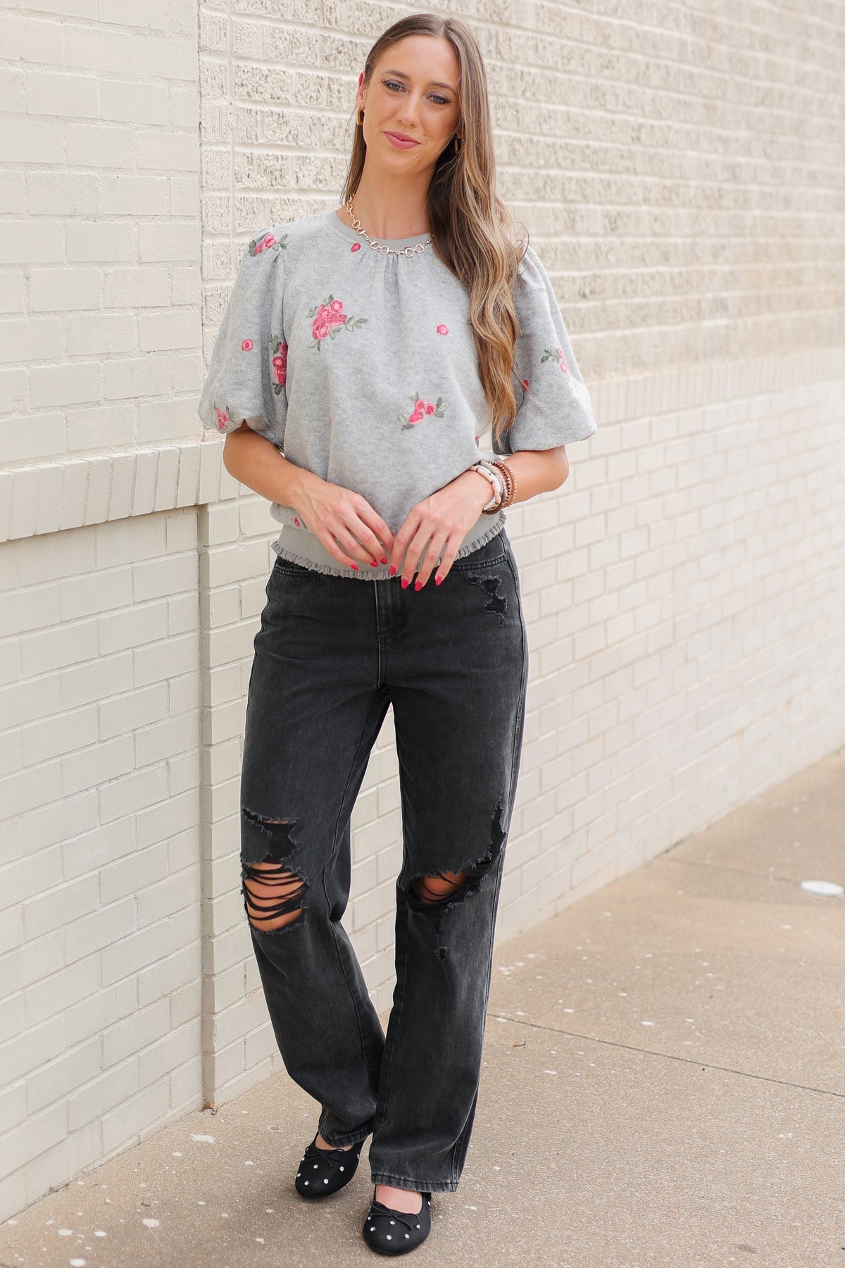 Blonde model wearing a heather grey sweater with rose patterns, black denim pants, and black ballet flats, standing outside. Southern House Clothing