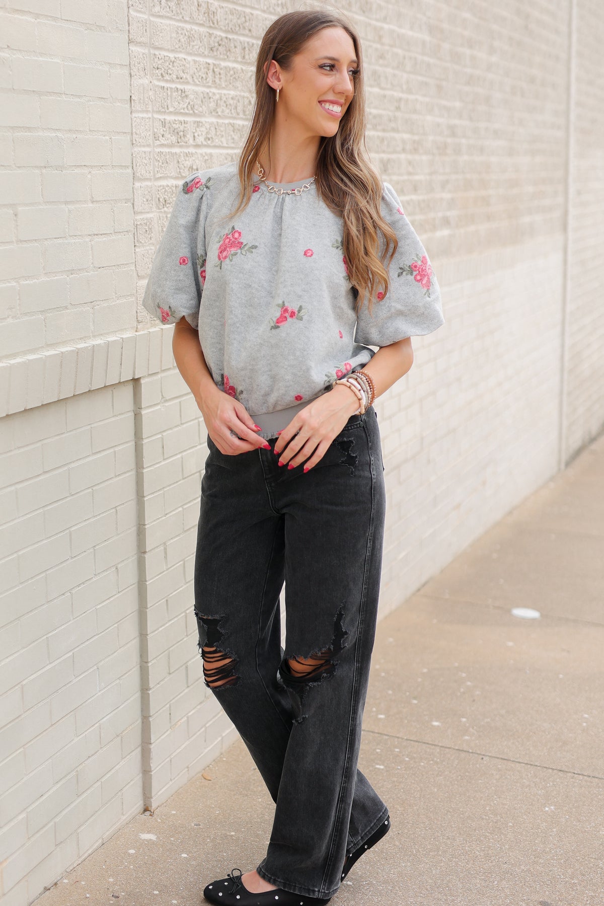 Blonde model wearing a heather grey sweater with rose patterns, black denim pants, and black ballet flats, standing outside. Southern House Clothing