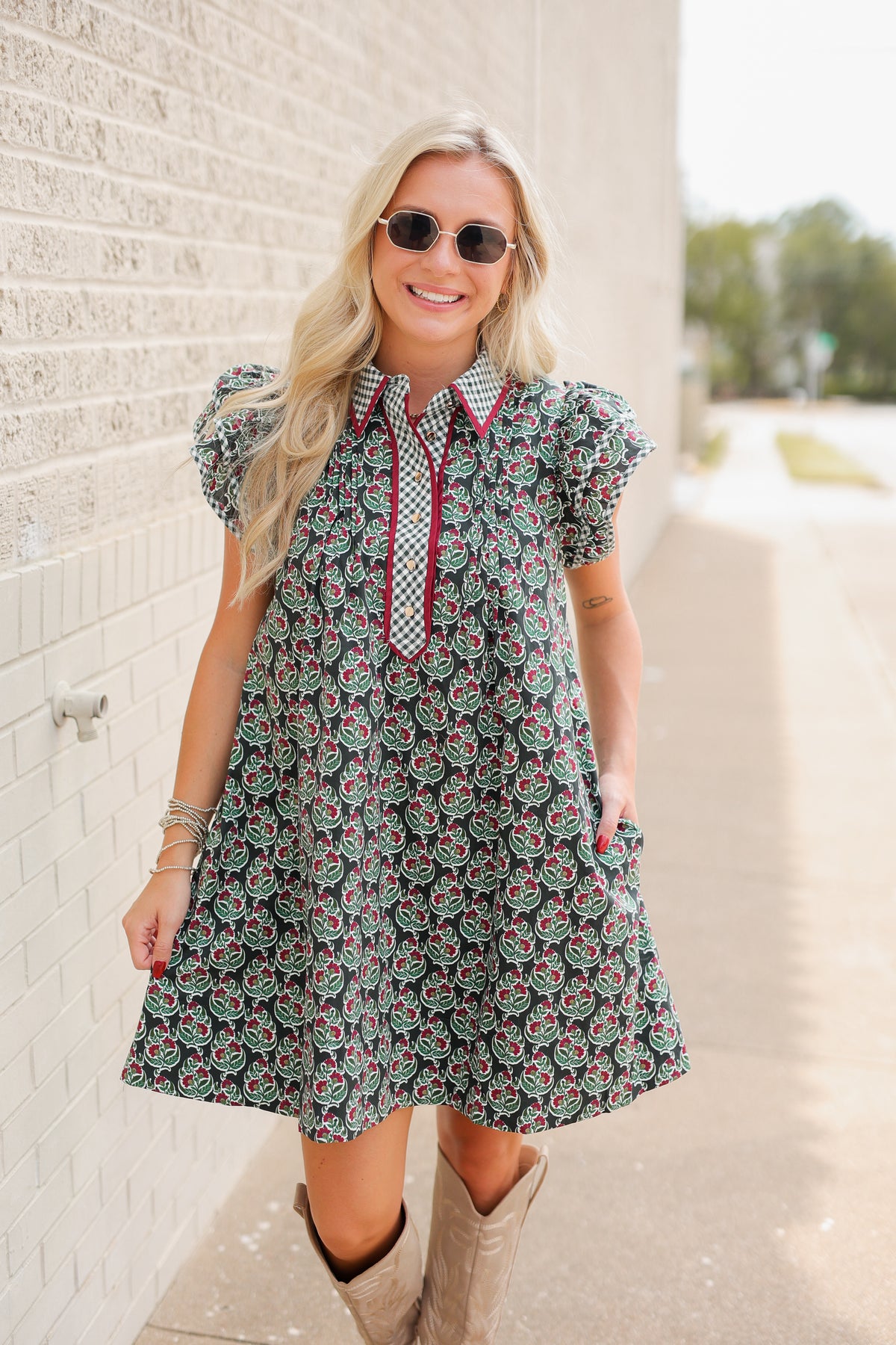 Blonde model standing outside wearing a green dress with red trim and brown cowboys boots. Southern House Clothing