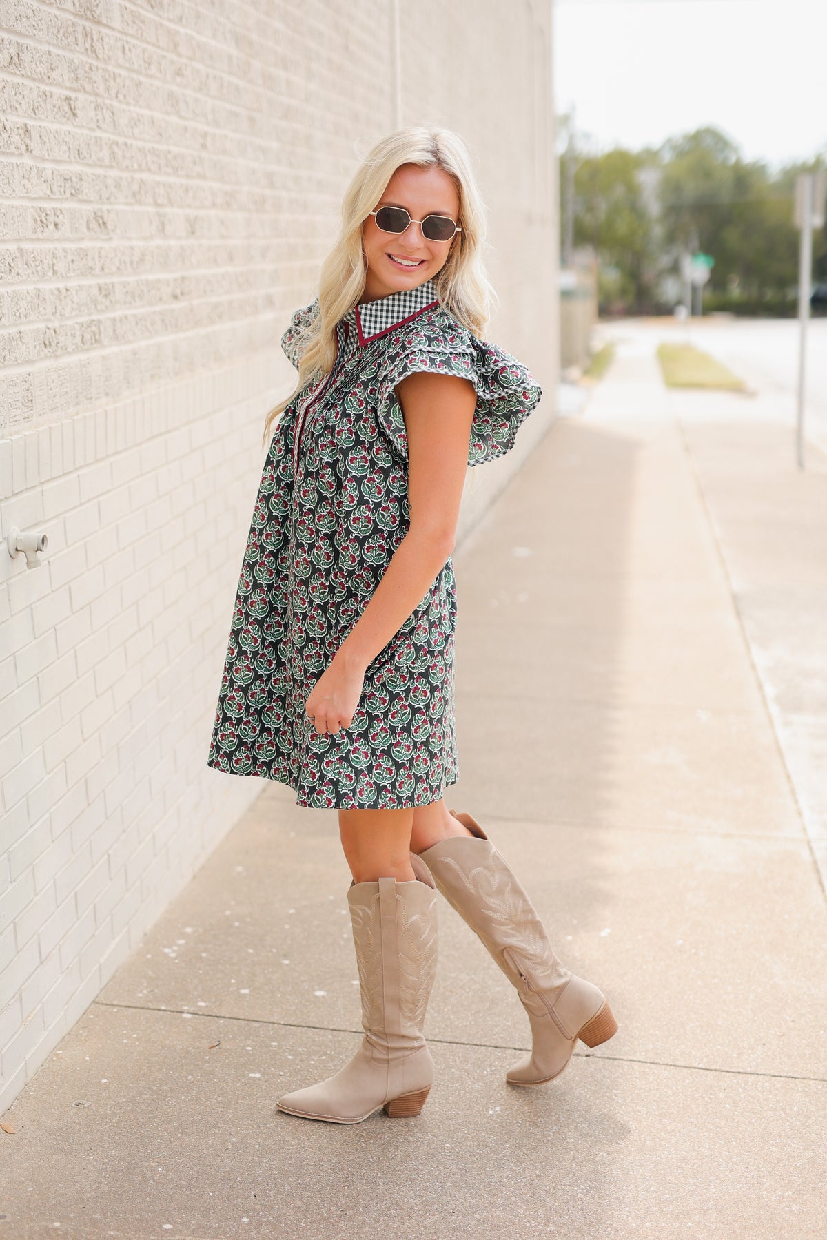 Blonde model standing outside wearing a green dress with red trim and brown cowboys boots. Southern House Clothing