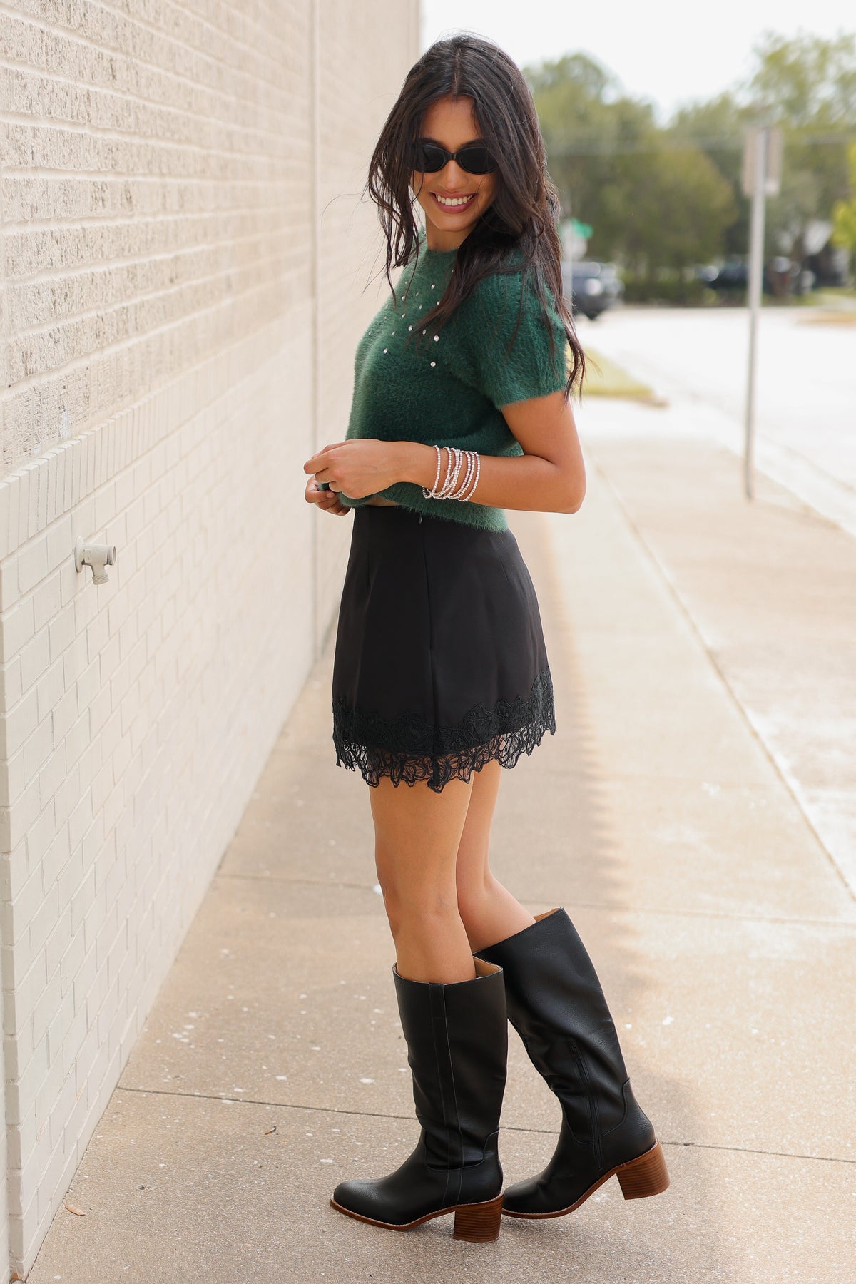 A brunnette model wearing the Liora Pearl Knit Top – Hunter Green, the Bellarosa skort in black and the harriett tall riding boots in black standing outside. Southern House Clothing