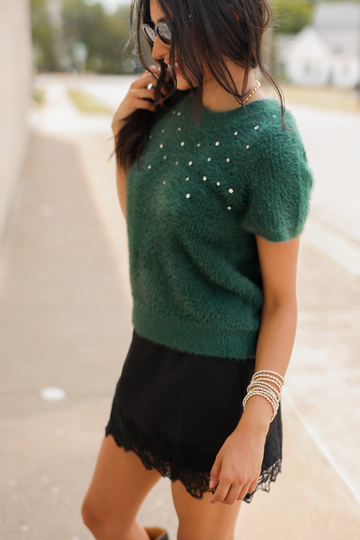 A brunnette model wearing the Liora Pearl Knit Top – Hunter Green, the Bellarosa skort in black and the harriett tall riding boots in black standing outside. Southern House Clothing