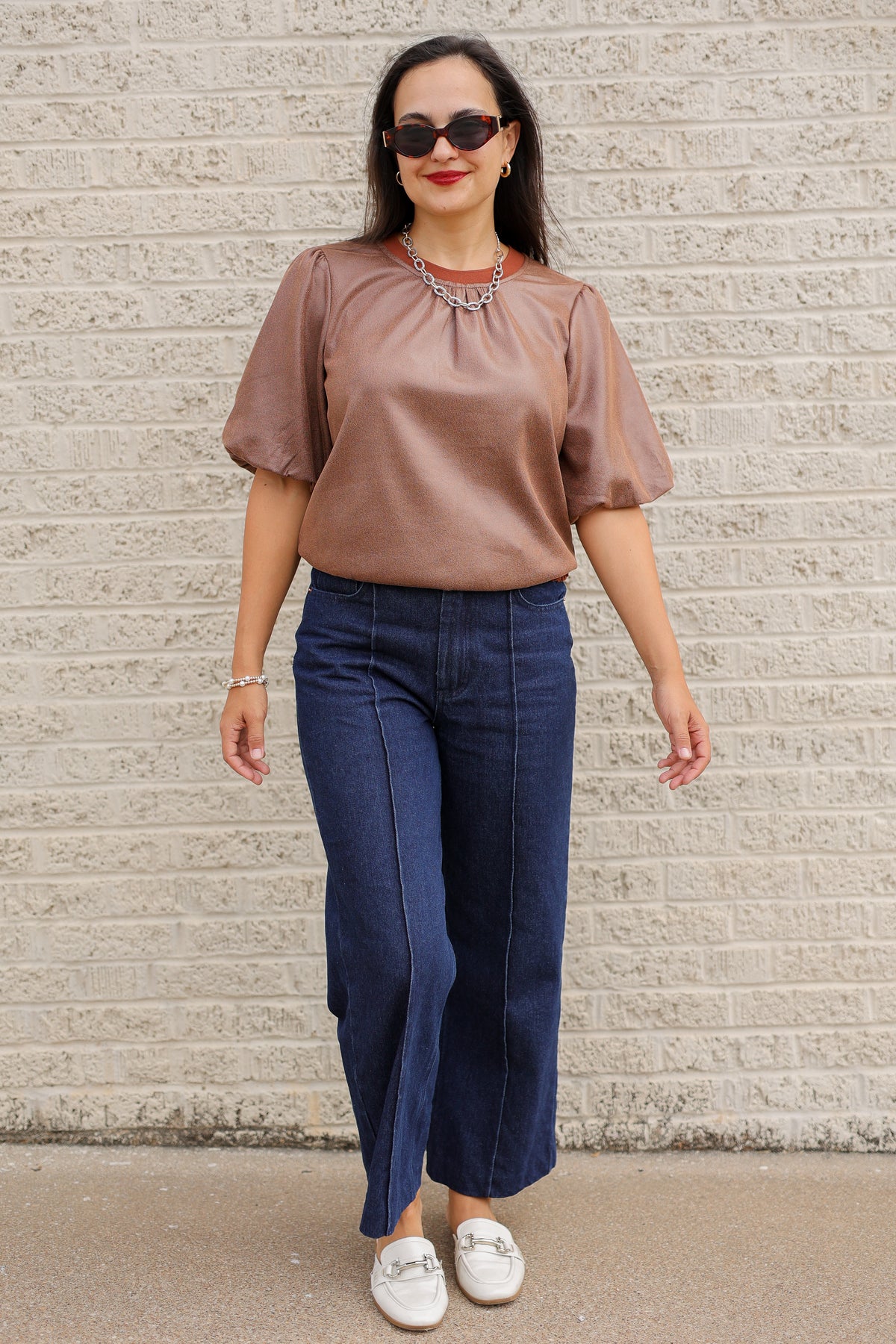 Brunette model standing outside wearing a brown blouse and denim jeans wearing black sunglasses.