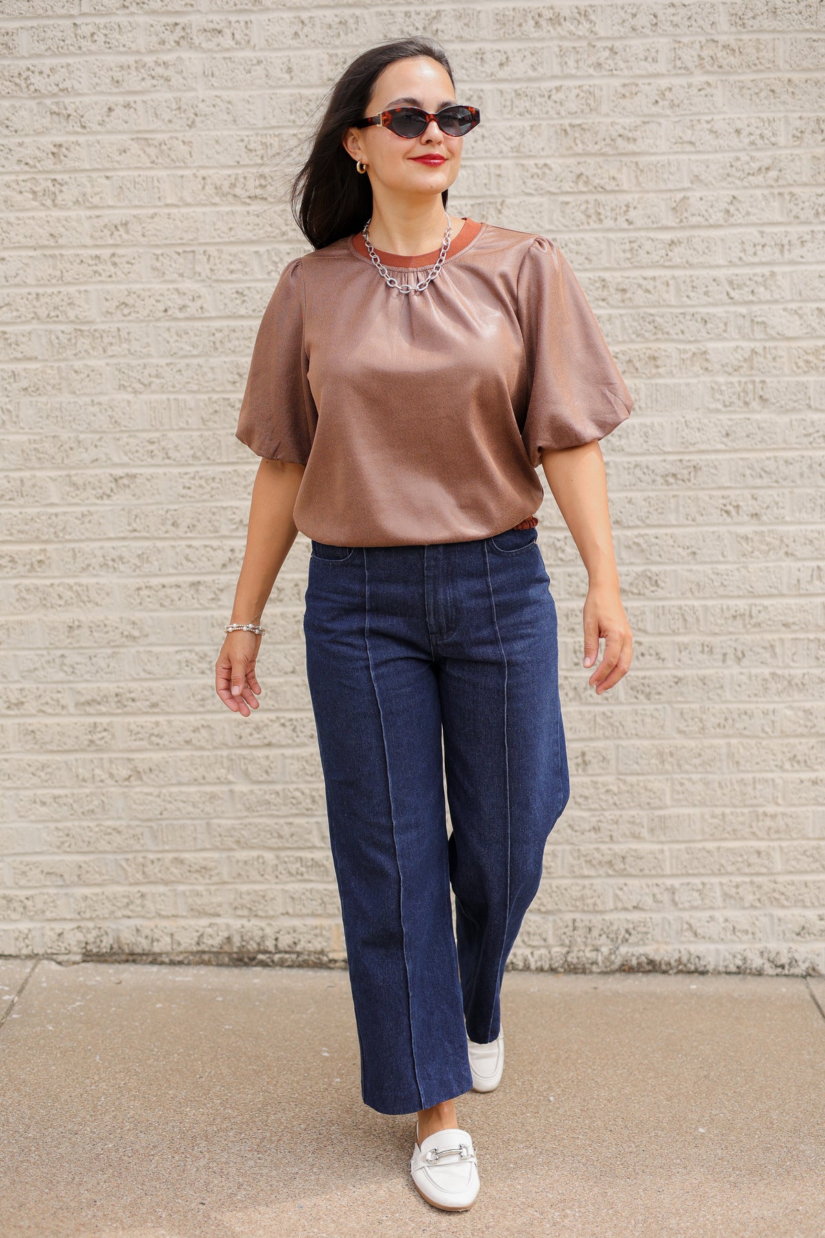 Brunette model standing outside wearing a brown blouse and denim jeans wearing black sunglasses.