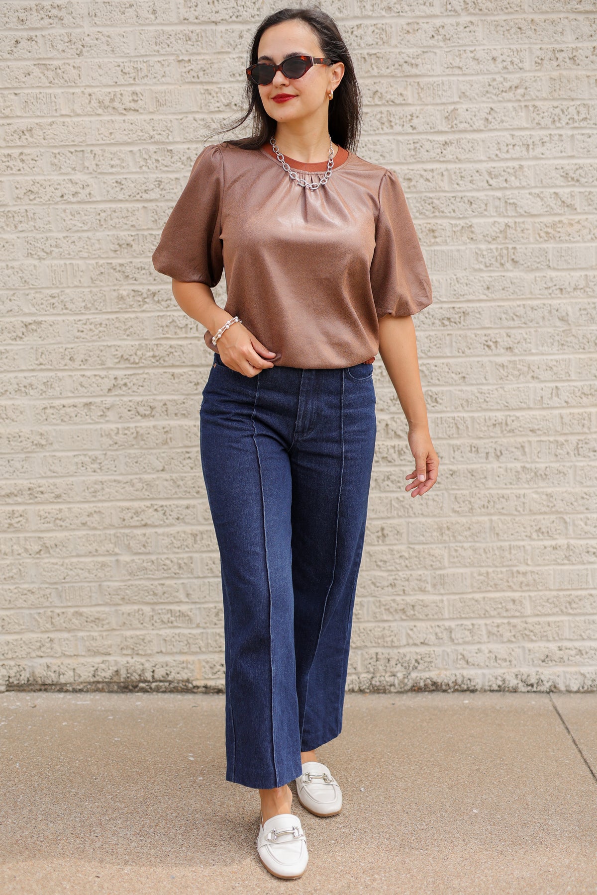 Brunette model standing outside wearing a brown blouse and denim jeans wearing black sunglasses.