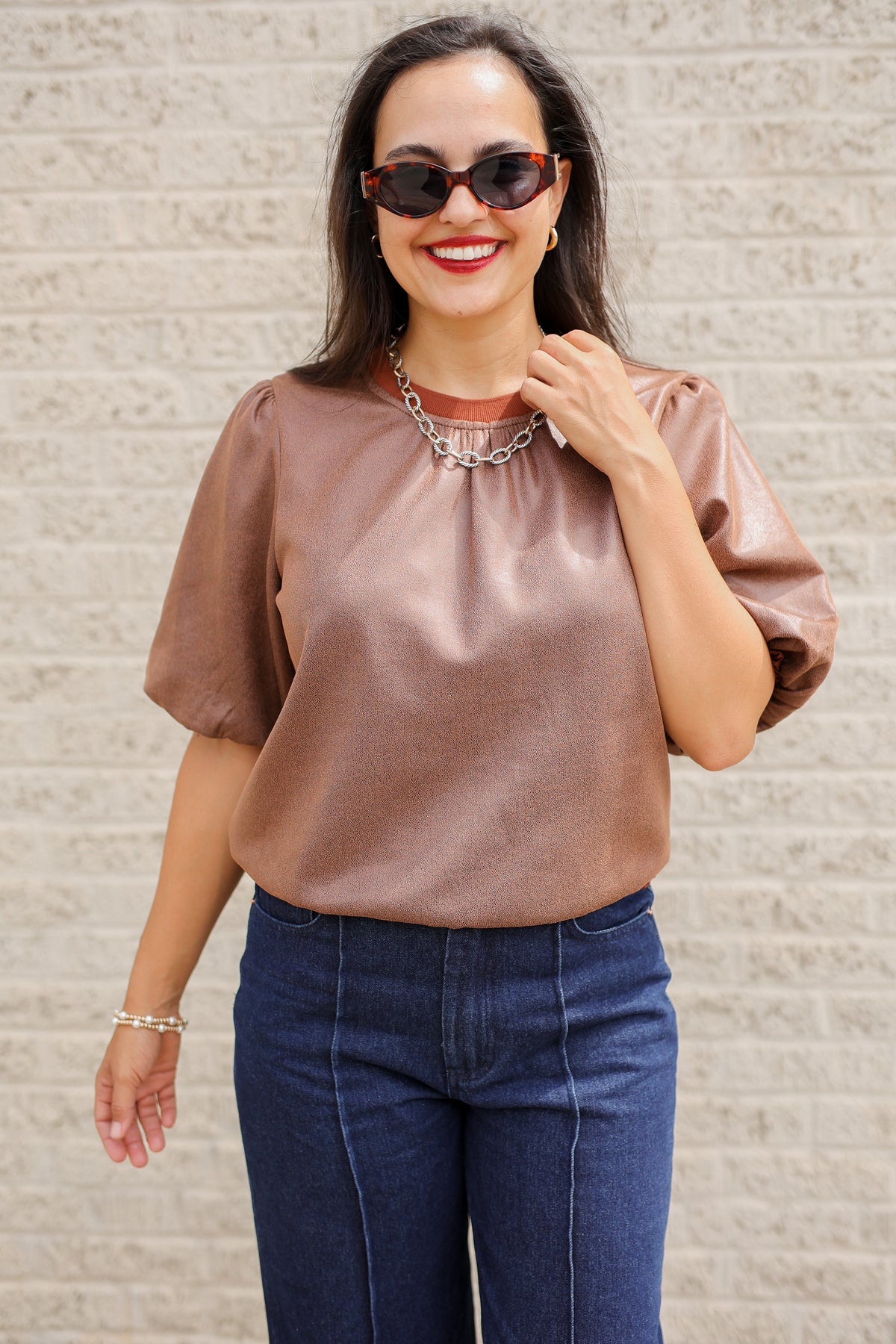 Brunette model standing outside wearing a brown blouse and denim jeans wearing black sunglasses.