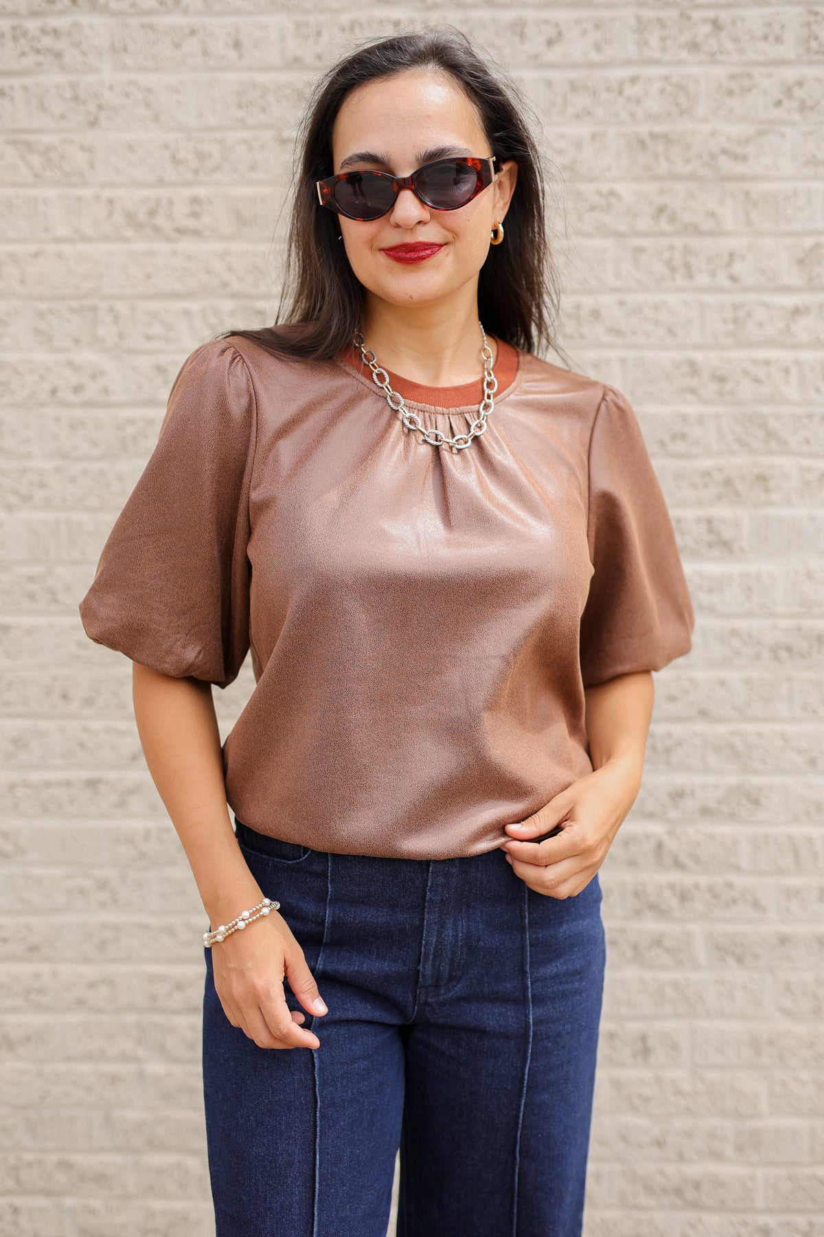 Brunette model standing outside wearing a brown blouse and denim jeans wearing black sunglasses.