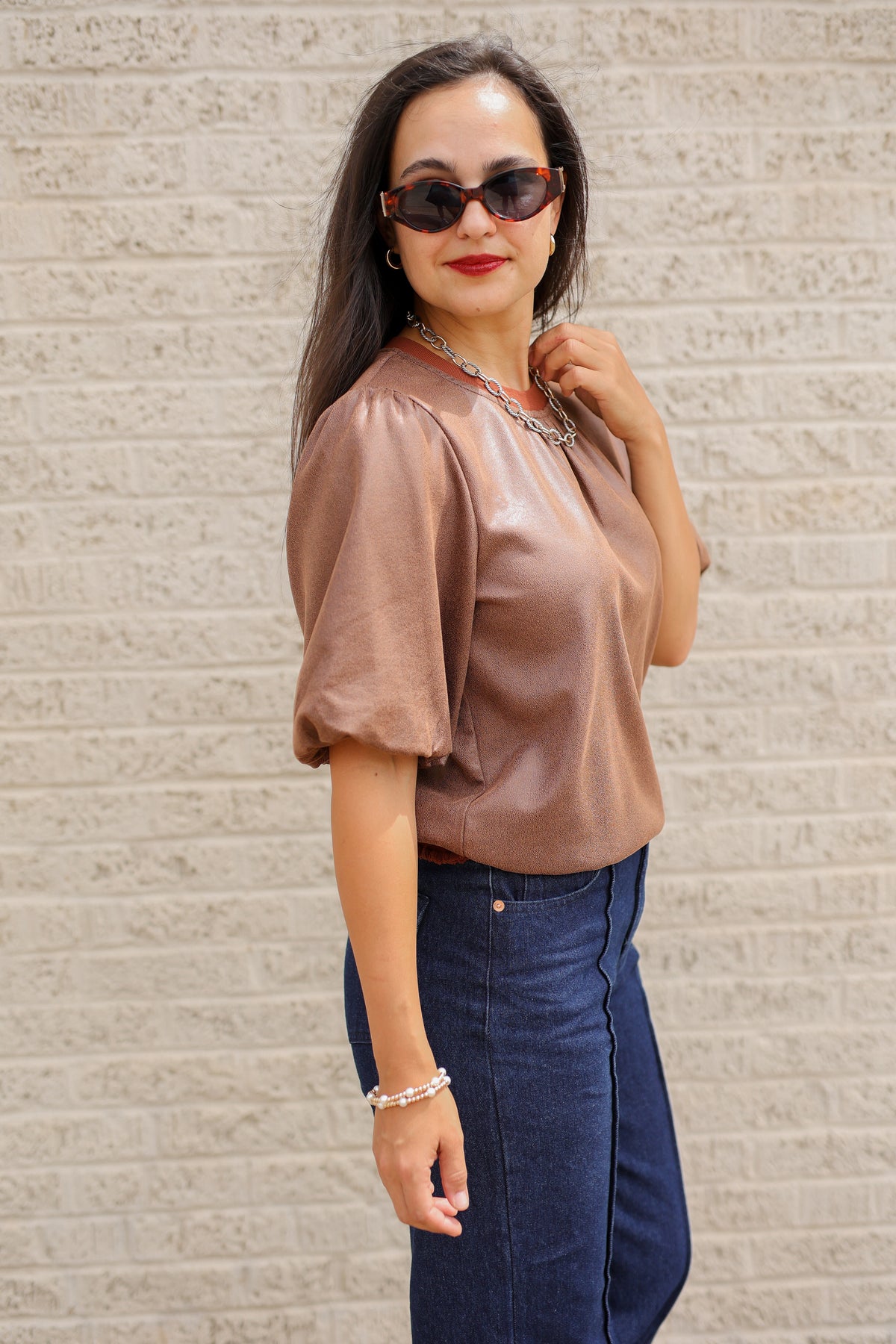 Brunette model standing outside wearing a brown blouse and denim jeans wearing black sunglasses.