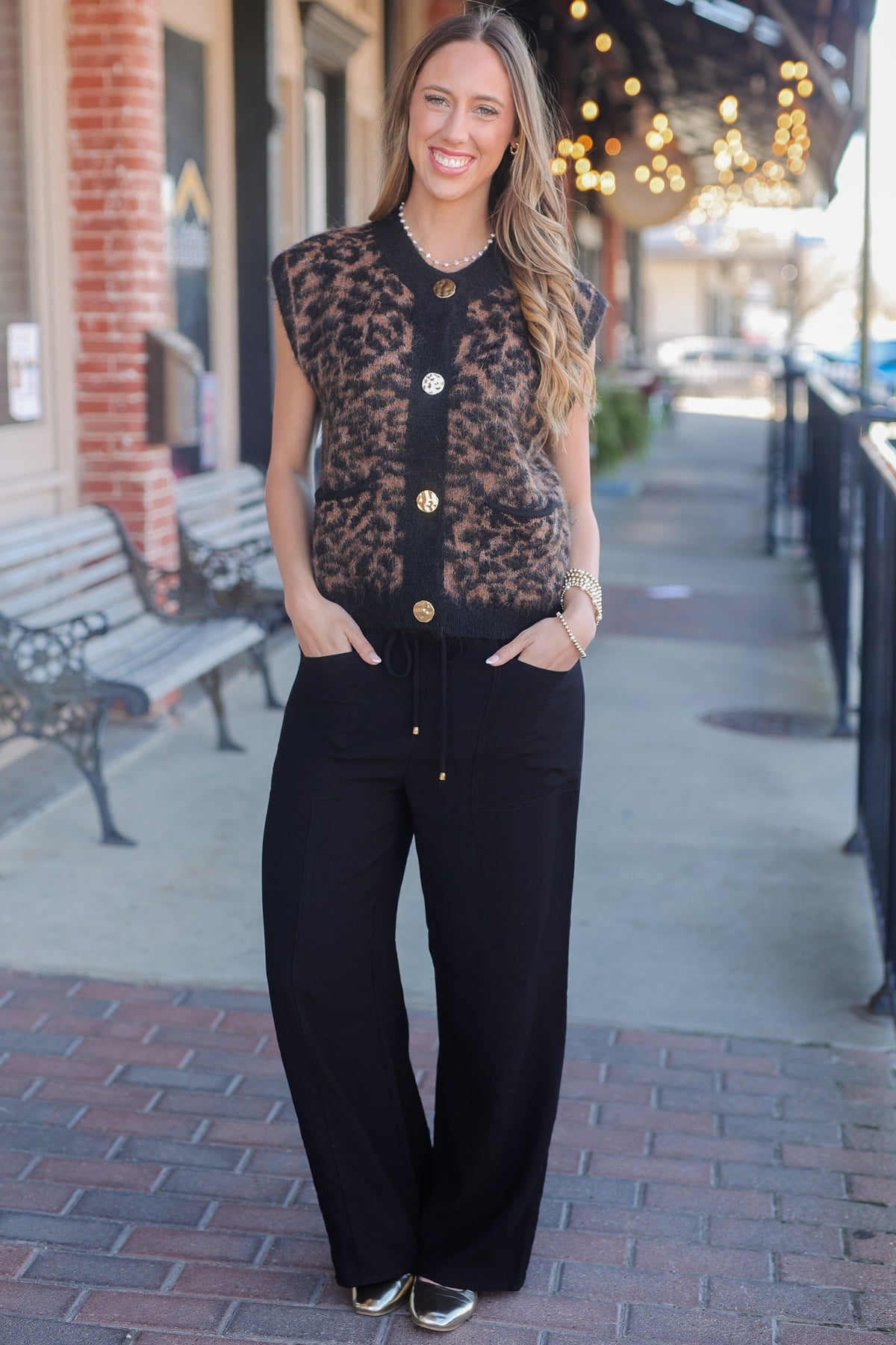 Woman wearing a black sleeveless top with gold buttons and black pants on a sidewalk.
