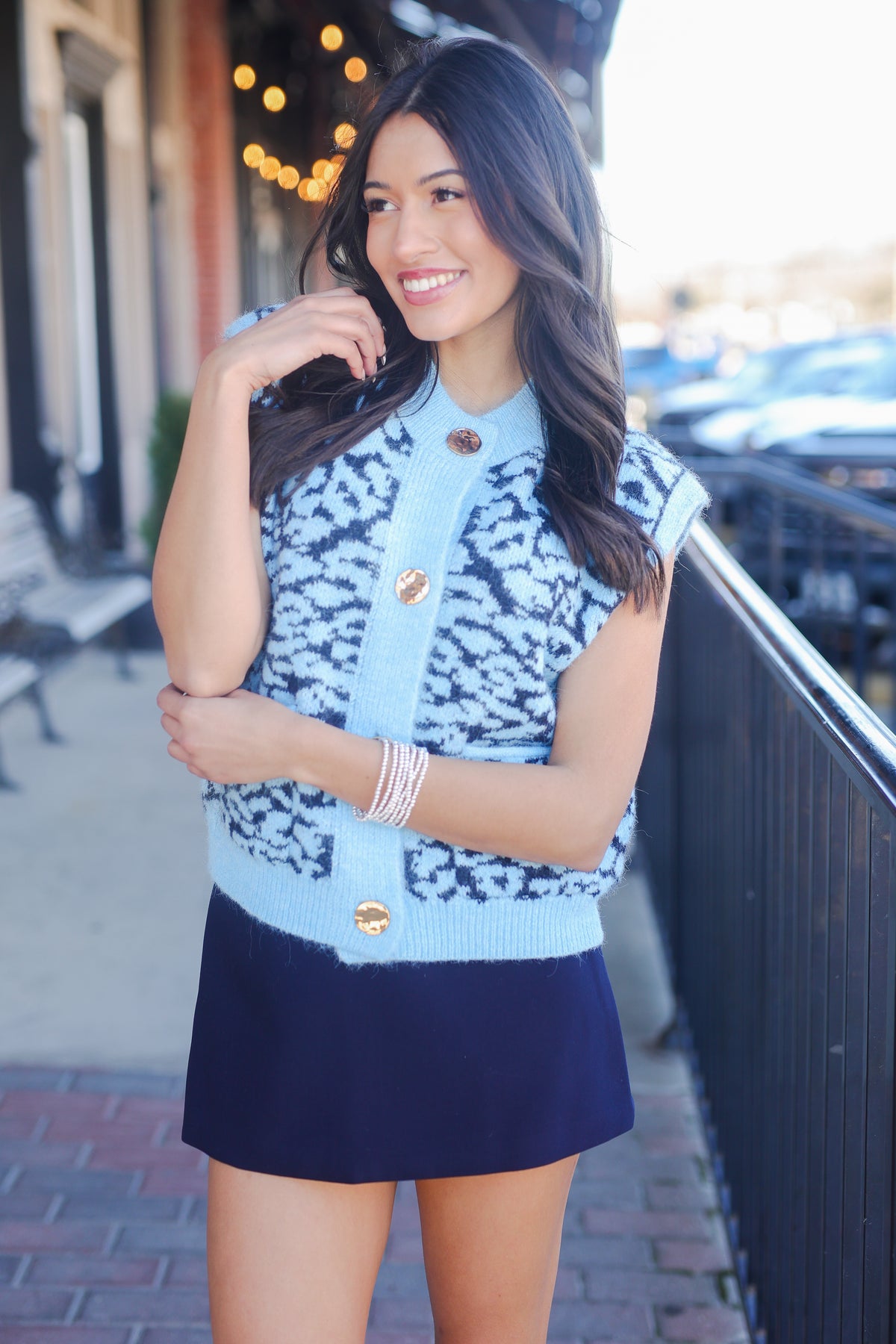 Woman wearing a blue patterned sleeveless top and navy skirt on a city street.
