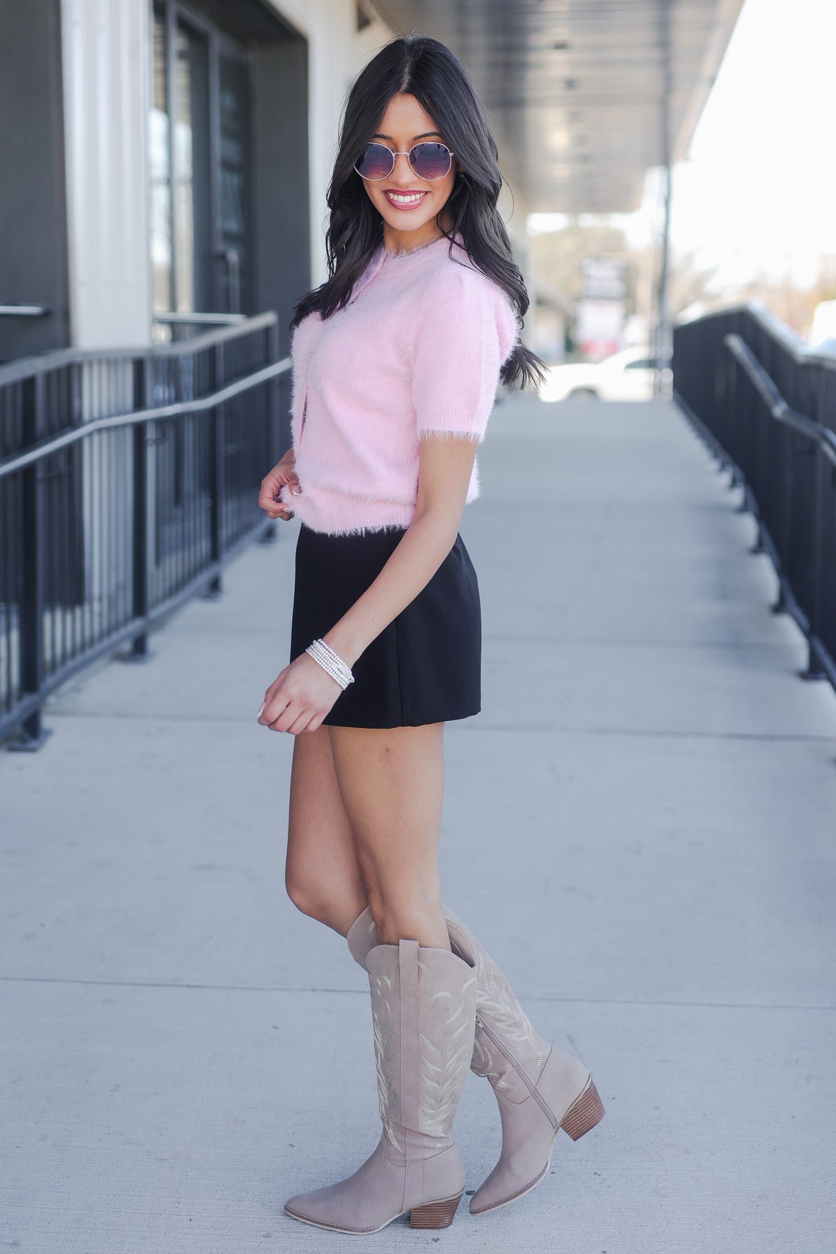 Woman wearing a pink sweater, black skirt, and beige knee-high boots on a sidewalk.