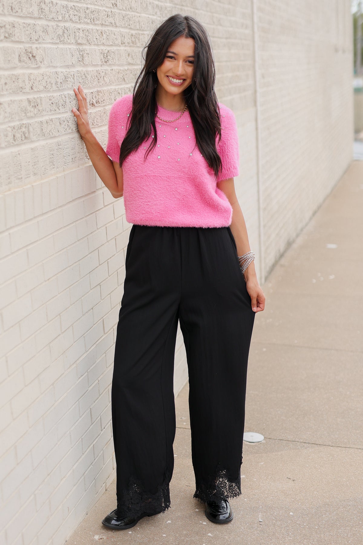 Brunette model wearing the Liora Pearl Knit Top –  Flamingo - and the Ellison pants in black with the toulouse flats on standing outside. Southern House Clothing