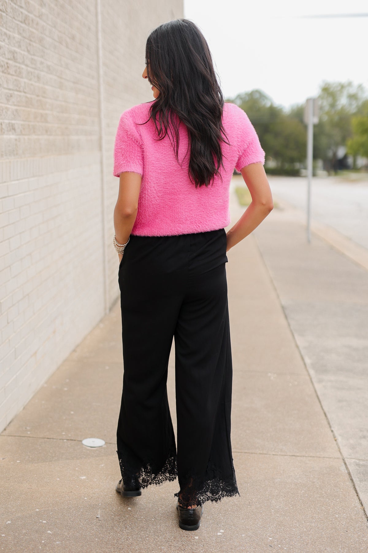 Brunette model wearing a pink sweater top and black pants with a lace detail standing outside. Southern House Clothing