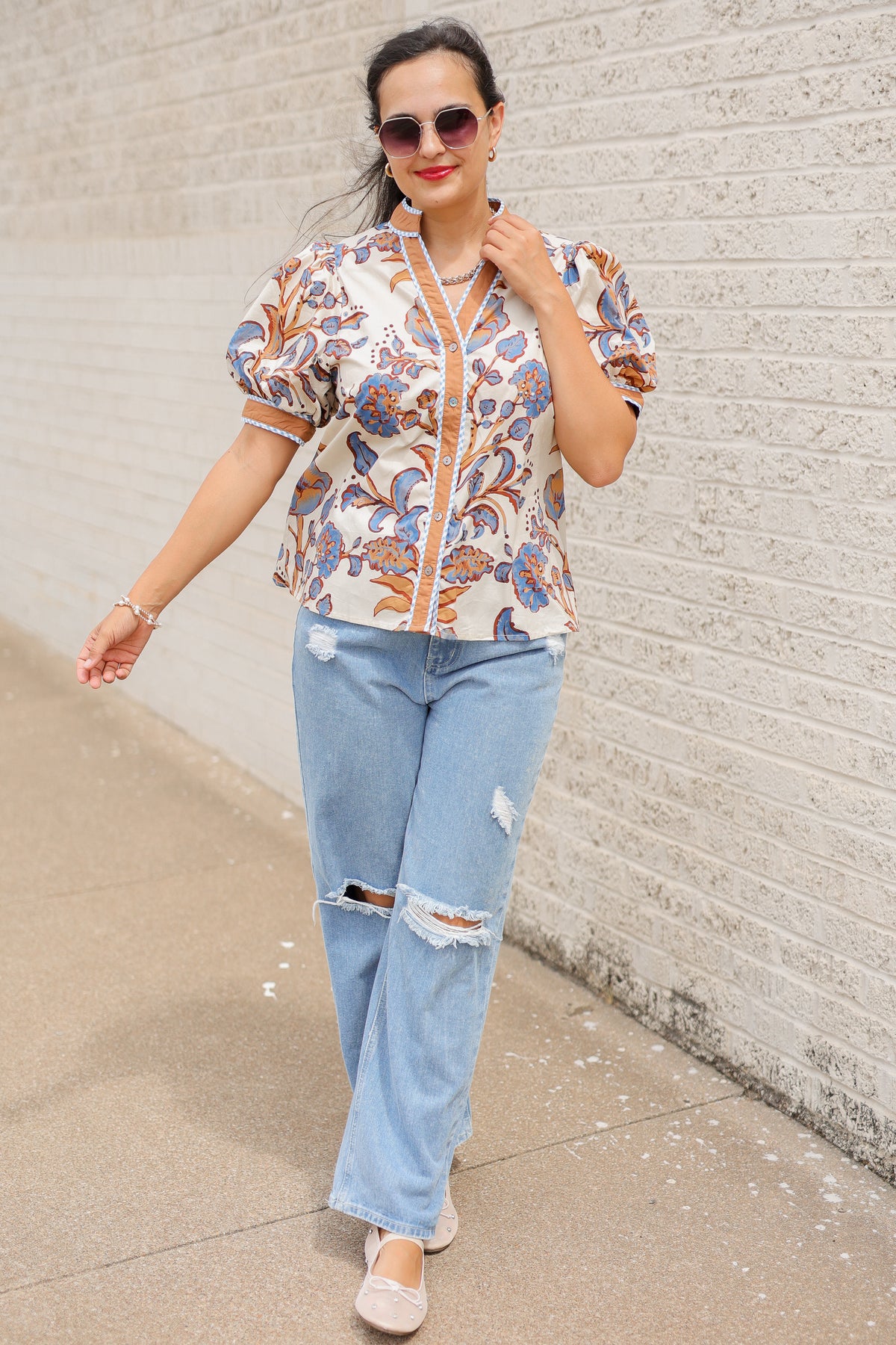 Brunette model wearing a ecru, tan, and blue floral blouse with sunglasses on and denim pants, standing outside. Southern House Clothing