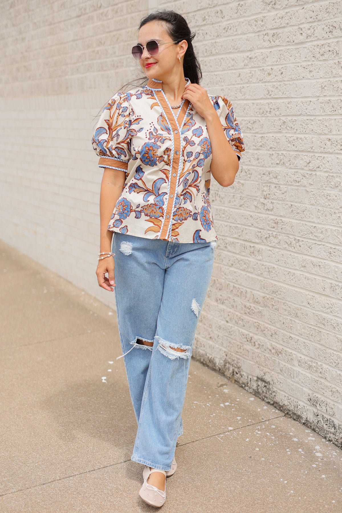 Brunette model wearing a ecru, tan, and blue floral blouse with sunglasses on and denim pants, standing outside. Southern House Clothing