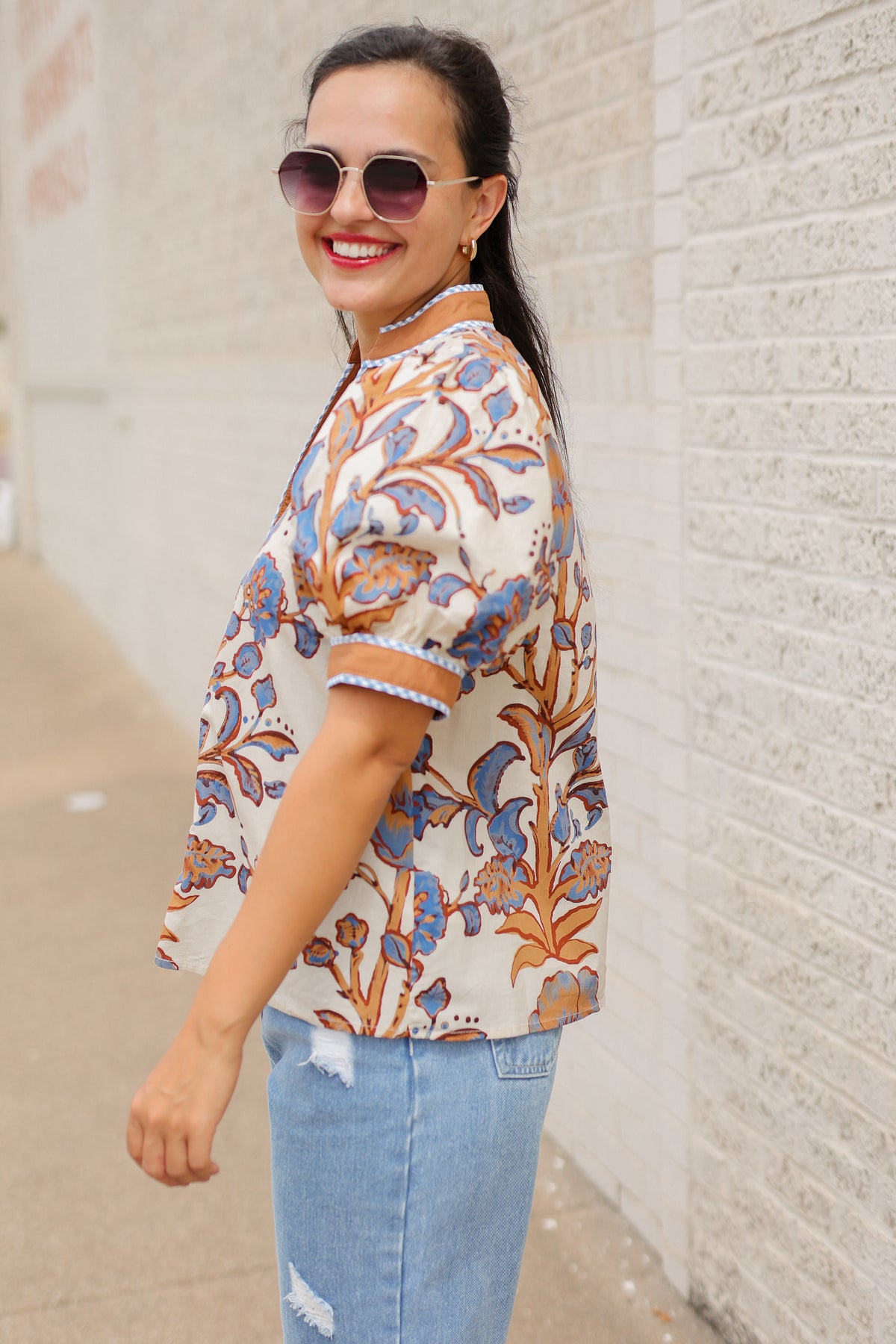 Brunette model wearing a ecru, tan, and blue floral blouse with sunglasses on and denim pants, standing outside. Southern House Clothing