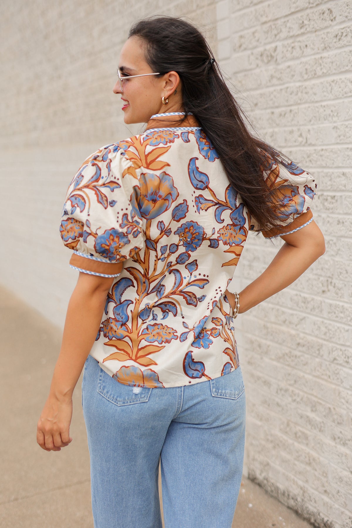 Brunette model wearing a ecru, tan, and blue floral blouse with sunglasses on and denim pants, standing outside. Southern House Clothing