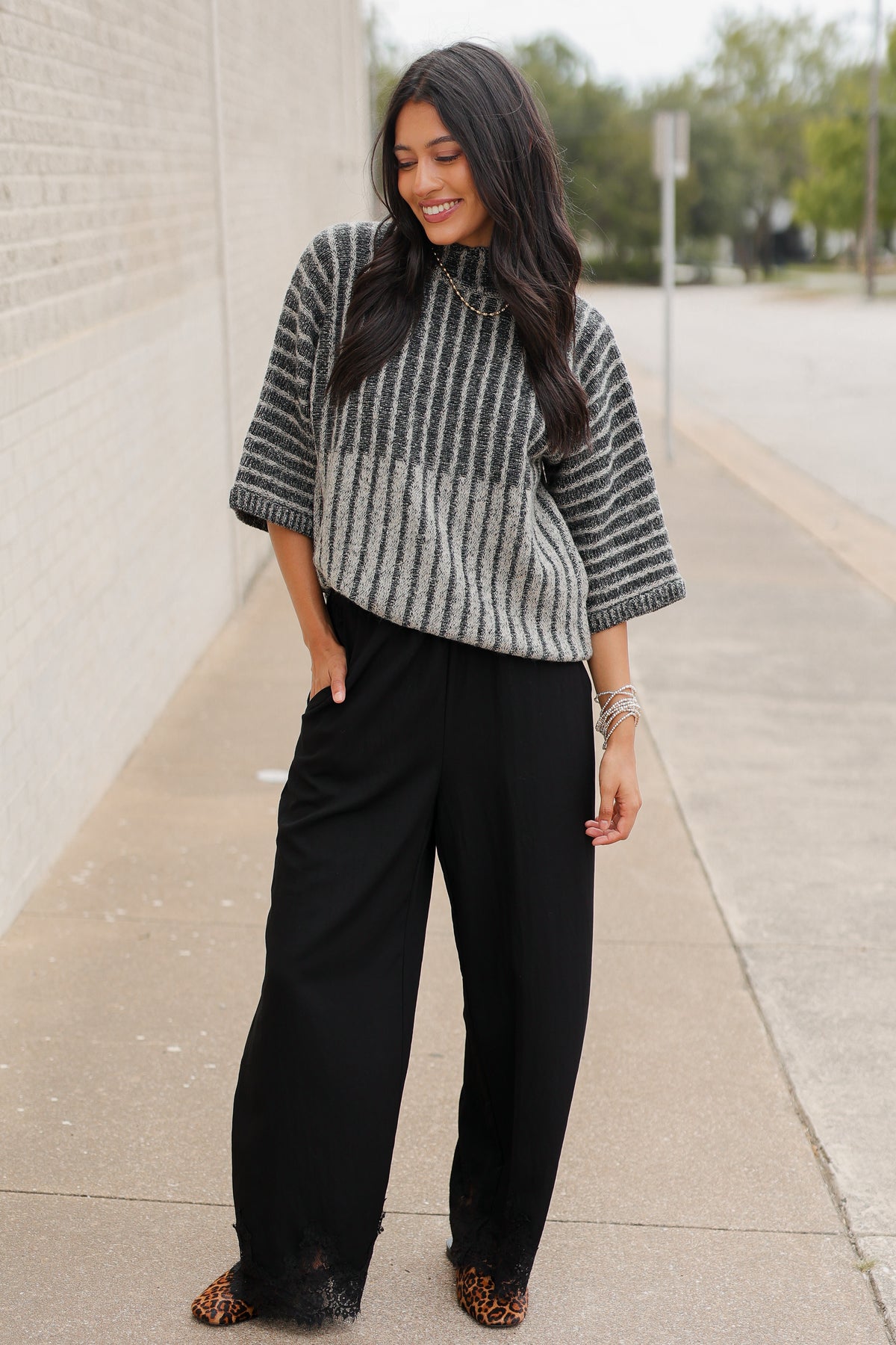 Brunette model wearing a black sweater top and black pants with a lace detail standing outside. Southern House Clothing