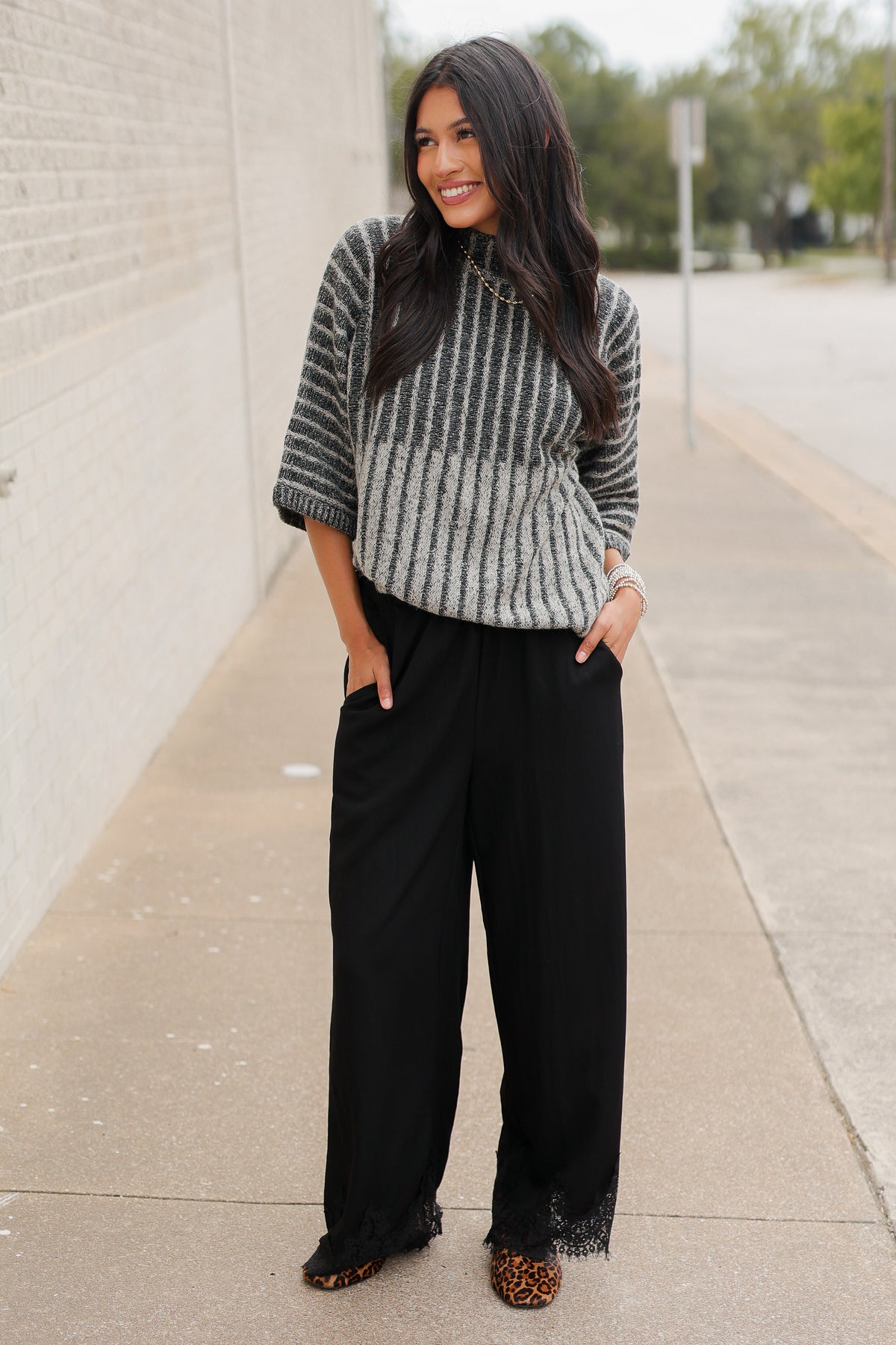 Brunette model wearing a black sweater top and black pants with a lace detail standing outside. Southern House Clothing
