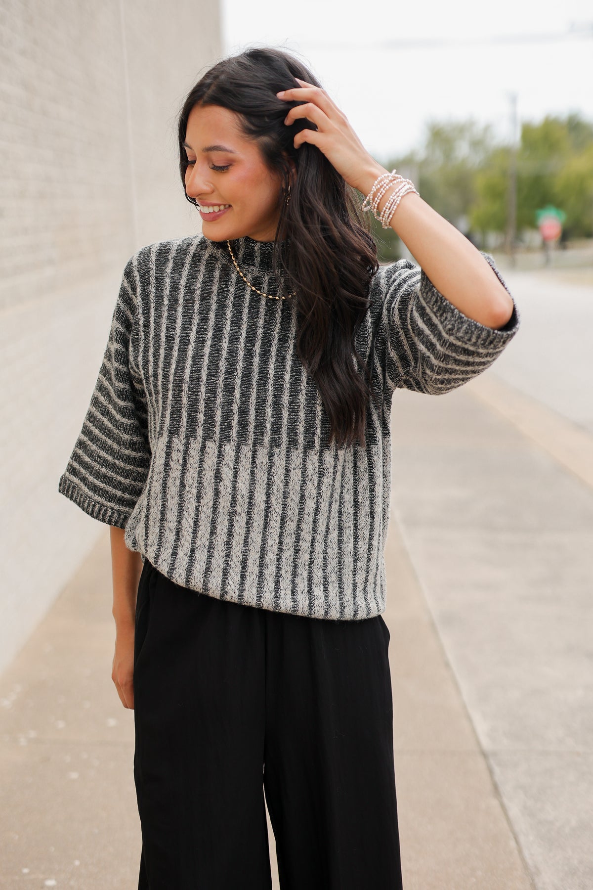 Brunette model wearing a black sweater top and black pants with a lace detail standing outside. Southern House Clothing