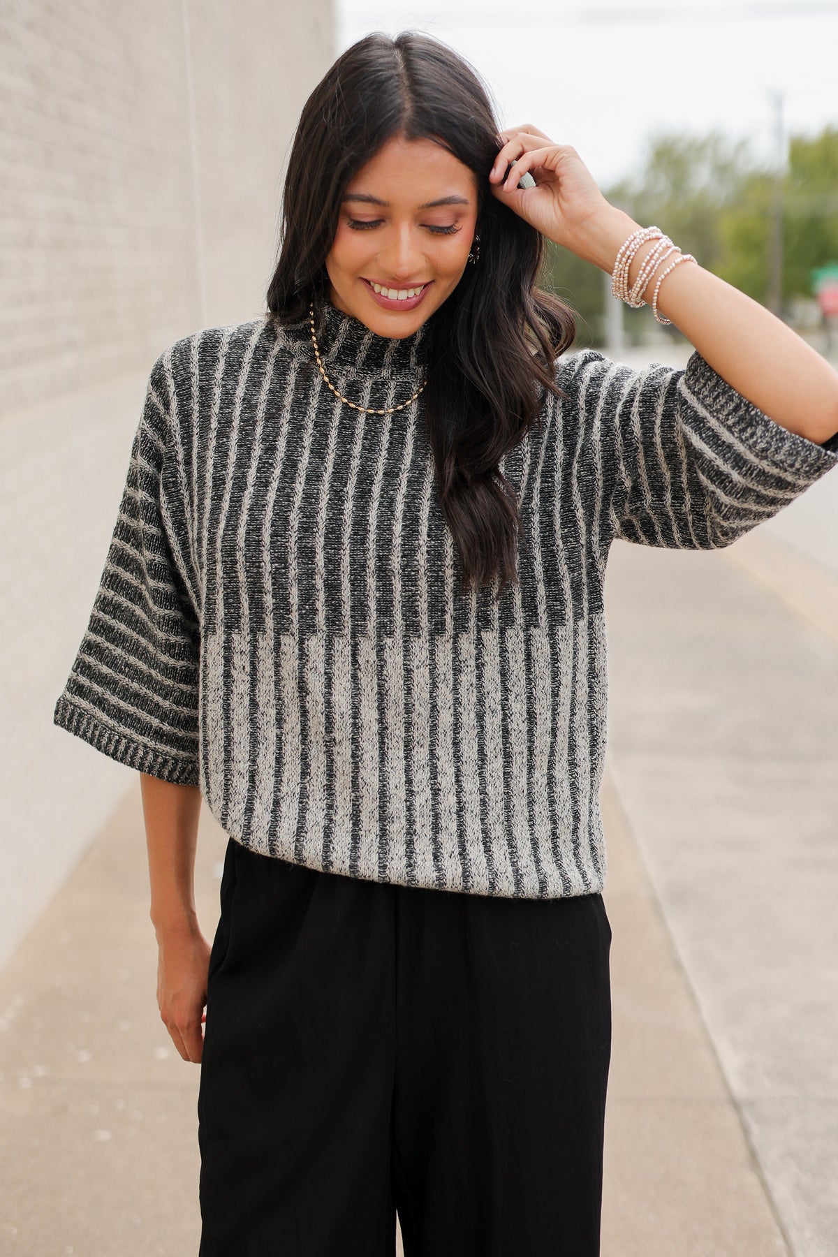 Brunette model wearing a black sweater top and black pants with a lace detail standing outside. Southern House Clothing