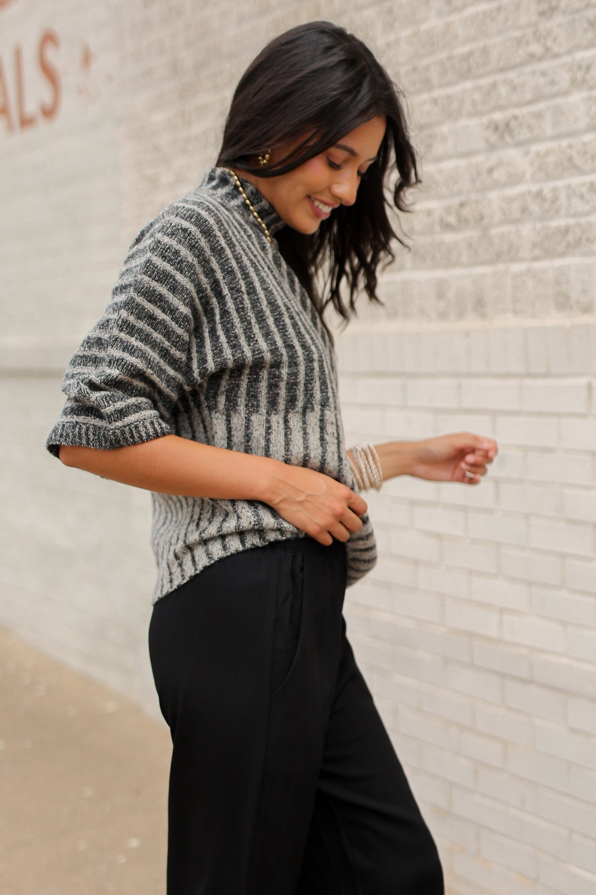 Brunette model wearing a black sweater top and black pants with a lace detail standing outside. Southern House Clothing