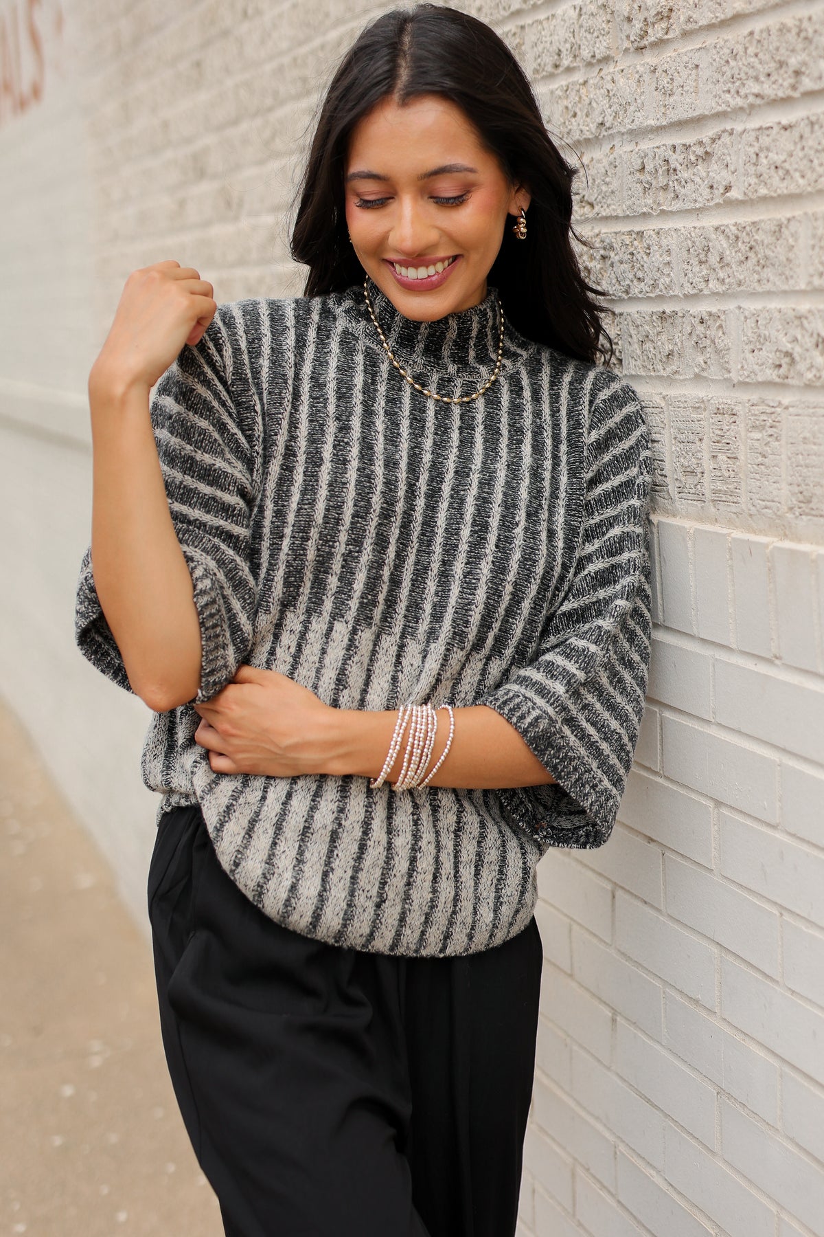 Brunette model wearing a black sweater top and black pants with a lace detail standing outside. Southern House Clothing