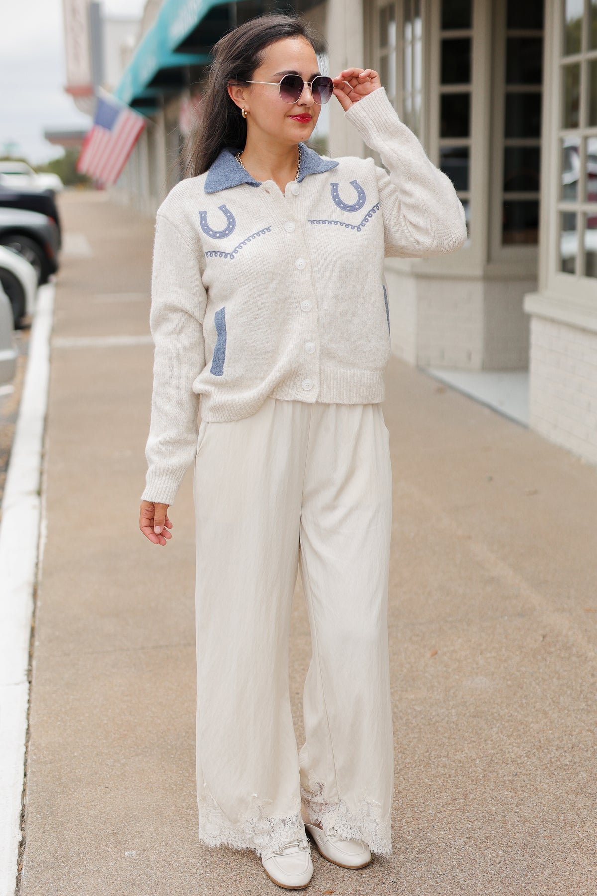 Brunette model wearing a Oatmeal knit Pecos Bridle Embroidered Cardigan with dusty blue collar, horseshoe embroidery, and front buttons, styled with cream trousers. Southern House Clothing