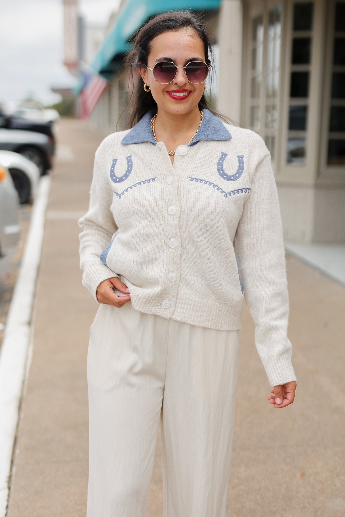 Brunette model wearing an Oatmeal knit Pecos Bridle Embroidered Cardigan with dusty blue collar, horseshoe embroidery, and front buttons, styled with cream trousers. Southern House Clothing