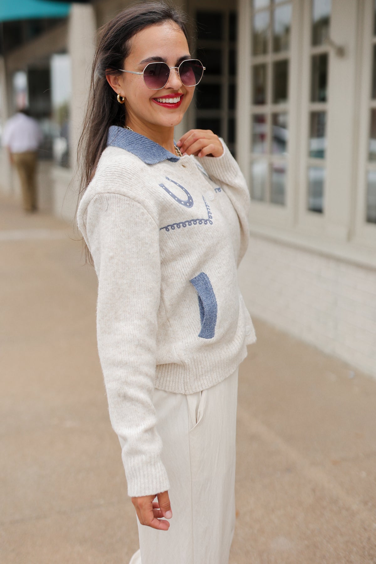 Brunette model wearing an Oatmeal knit Pecos Bridle Embroidered Cardigan with dusty blue collar, horseshoe embroidery, and front buttons, styled with cream trousers. Southern House Clothing