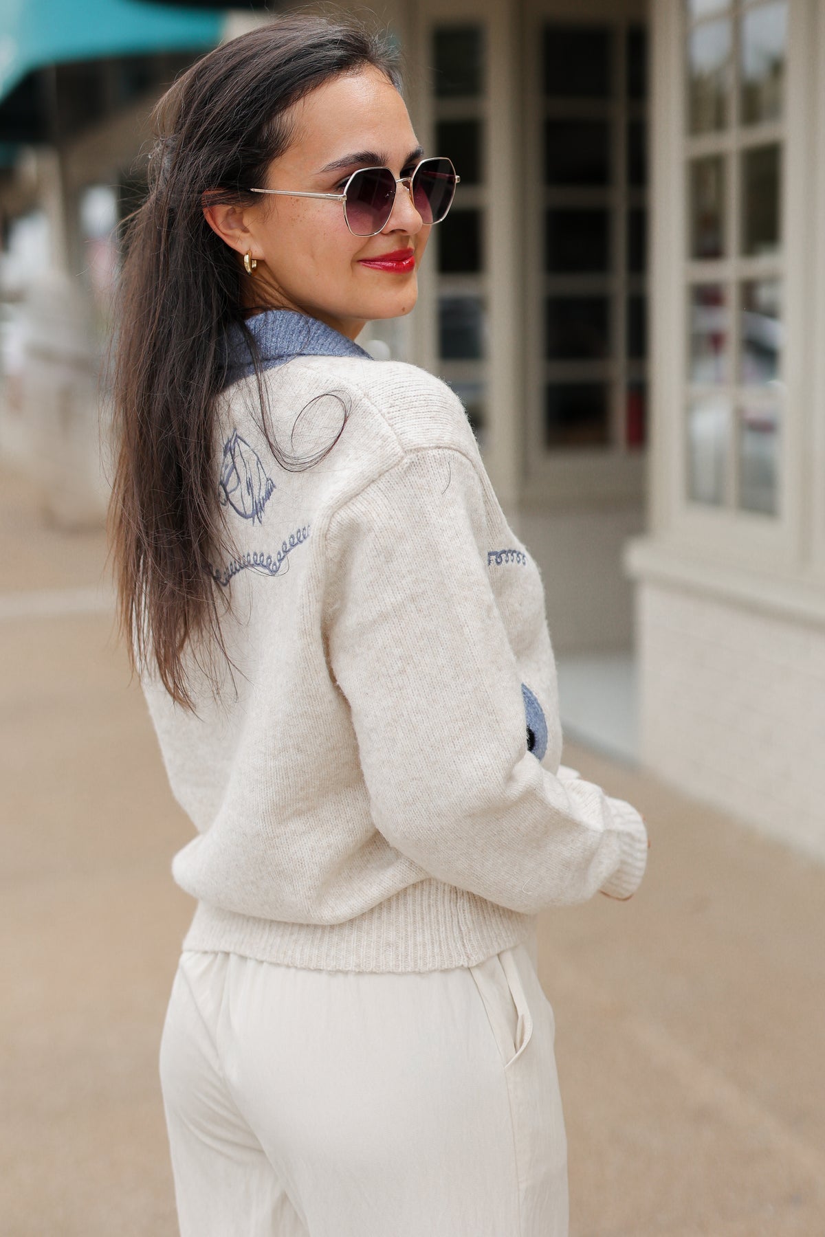 Brunette model wearing an Oatmeal knit Pecos Bridle Embroidered Cardigan with dusty blue collar, horseshoe embroidery, and front buttons, styled with cream trousers. Southern House Clothing
