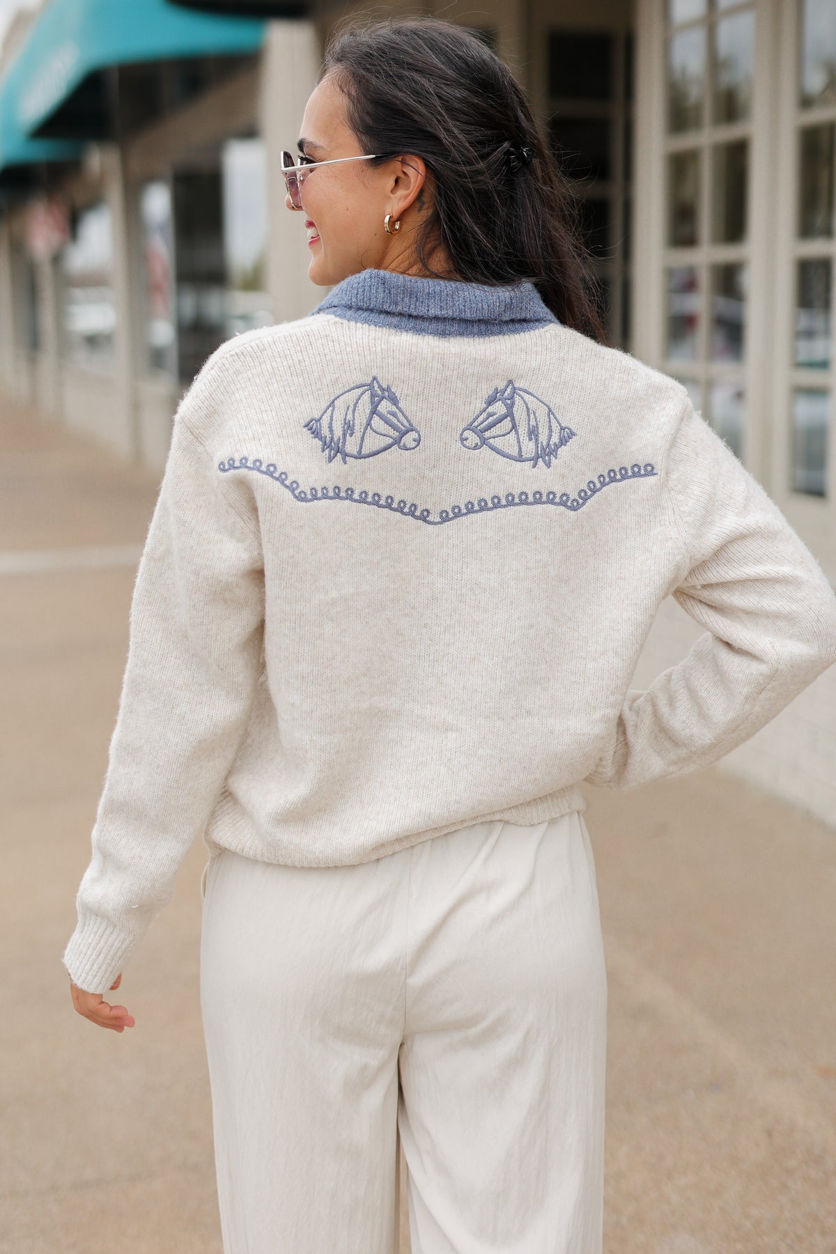 Brunette model wearing an Oatmeal knit Pecos Bridle Embroidered Cardigan with dusty blue collar, horseshoe embroidery, and front buttons, styled with cream trousers. Southern House Clothing